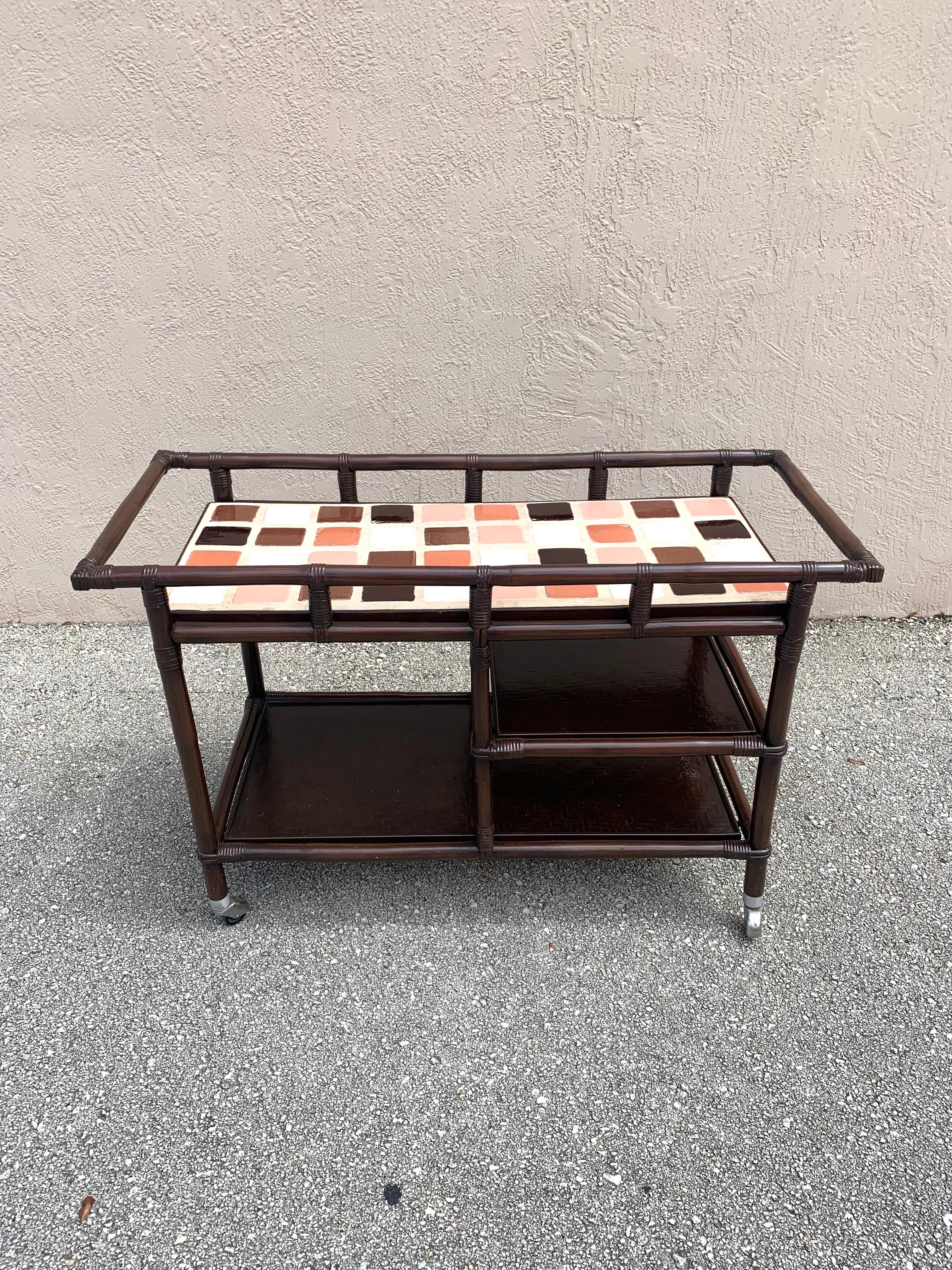 Rattan bar cart designed by John Wisner. Manufactured by Ficks Reed. Made of wood, cane and rattan. With a brown lacquer finish and a tile top of beautiful earth tones. Lattice bamboo work on the top of the shelves underneath. Circa 1950s. 

