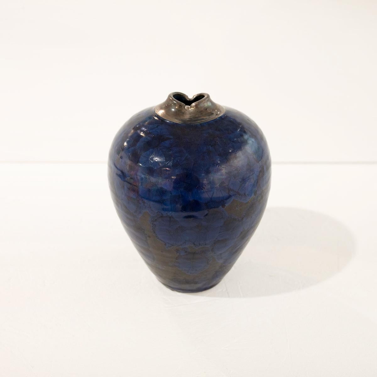 This small glazed porcelain sculptural vessel by Jon Puzzuoli features a blue palette with a crystalline glaze  a silver luster neck. The artist's stamp is located at the base of the vessel.

Crystalline glazes are special ceramic glazes in which