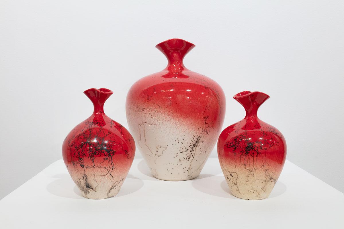 This ceramic vessel sculpture by Jon Puzzuoli is made with glazed porcelain. It has a vibrant red and cream palette, with a round body and a fluted neck. The piece has a raku finish, creating small black embellishments on the surface of the piece.