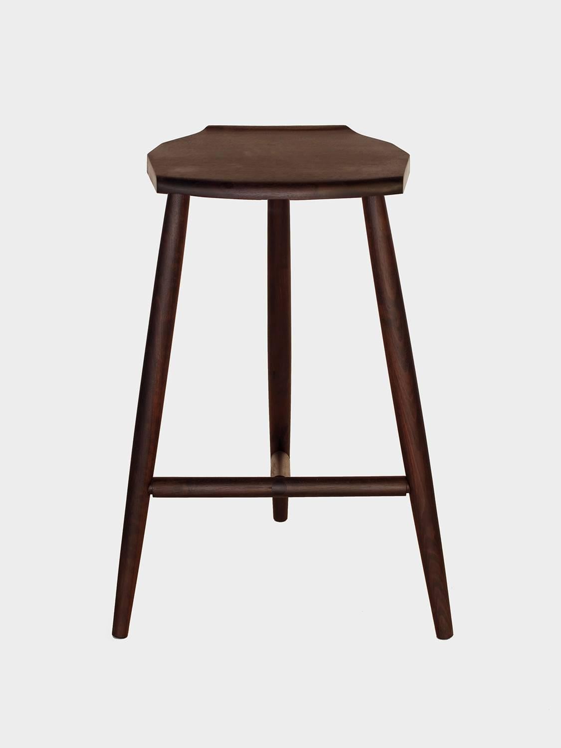 New York Heartwoods' ebonized walnut solid wood counter-height Jordan Stool is influenced by Shaker and Mid-Century design; created to be comfortable, light and easy to move; and features three turned legs, a unique hand-carved faceted seat, and