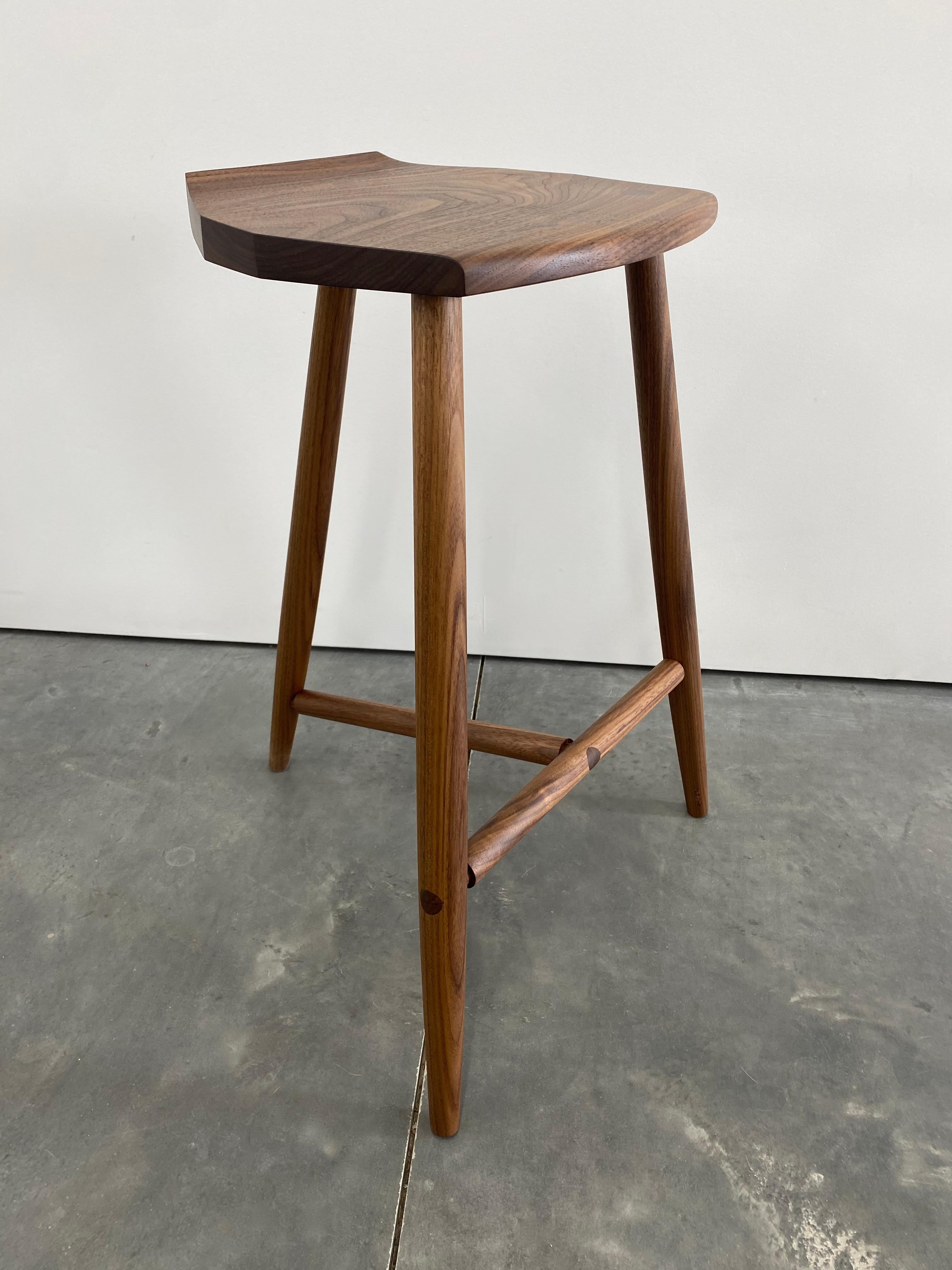 New York Heartwoods' walnut solid wood counter-height Jordan Stool is influenced by Shaker and Mid-Century design; created to be comfortable, light and easy to move; and features three turned legs, a unique hand-carved faceted seat, and wedged