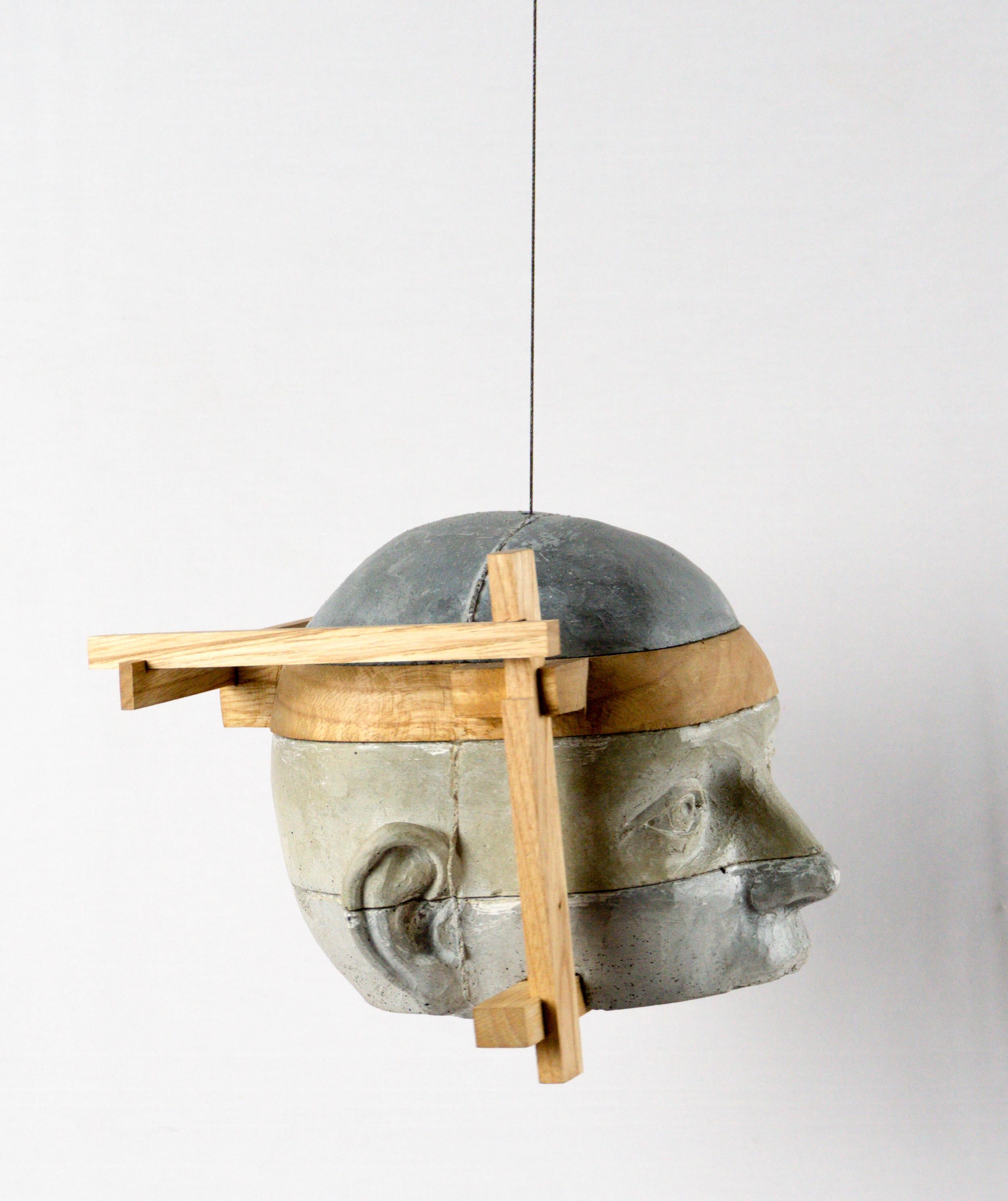 This architectural hanging cement head sculpture is defined by a distinctive lateral chestnut wood framework that surrounds the form like an external armature. In Project 23, the structure shifts outward from the head itself, creating a sense of