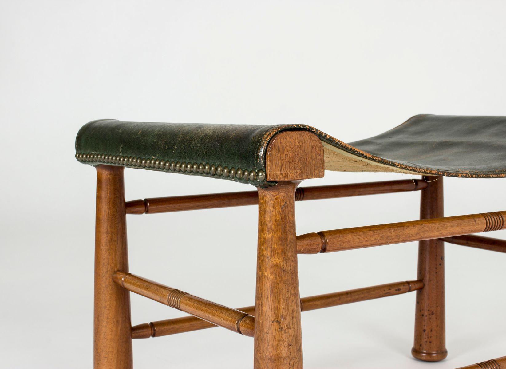 The stool Model 972 was created in 1941 by Swedish designer Josef Frank (1885-1967) for manufacture Svenskt Tenn, Stockholm. 
It is made of mahogany carved with linear decoration. The sling dark green leather seat is enhanced with brass nails to