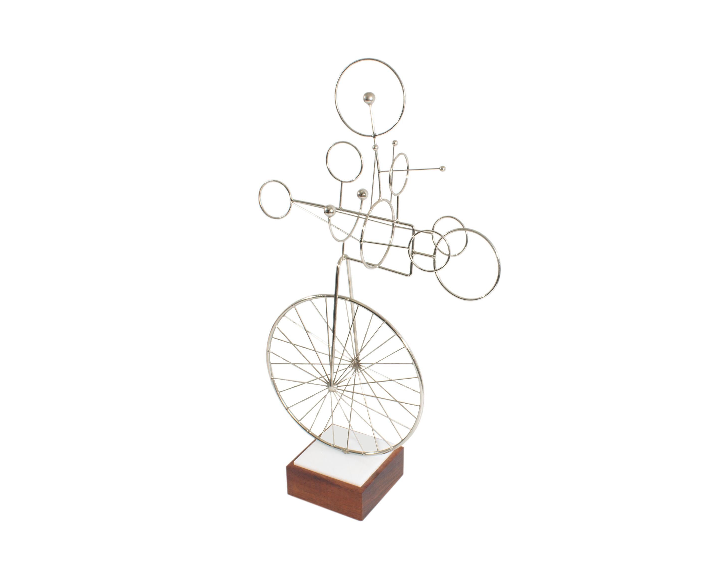 A metal bicycle sculpture by American artist Joseph Burlini (born 1937). Made in 1978, this sculpture is composed of multiple rods and circular chrome pieces forming an abstract bicycle attached to a white acrylic and wood base. The base is signed