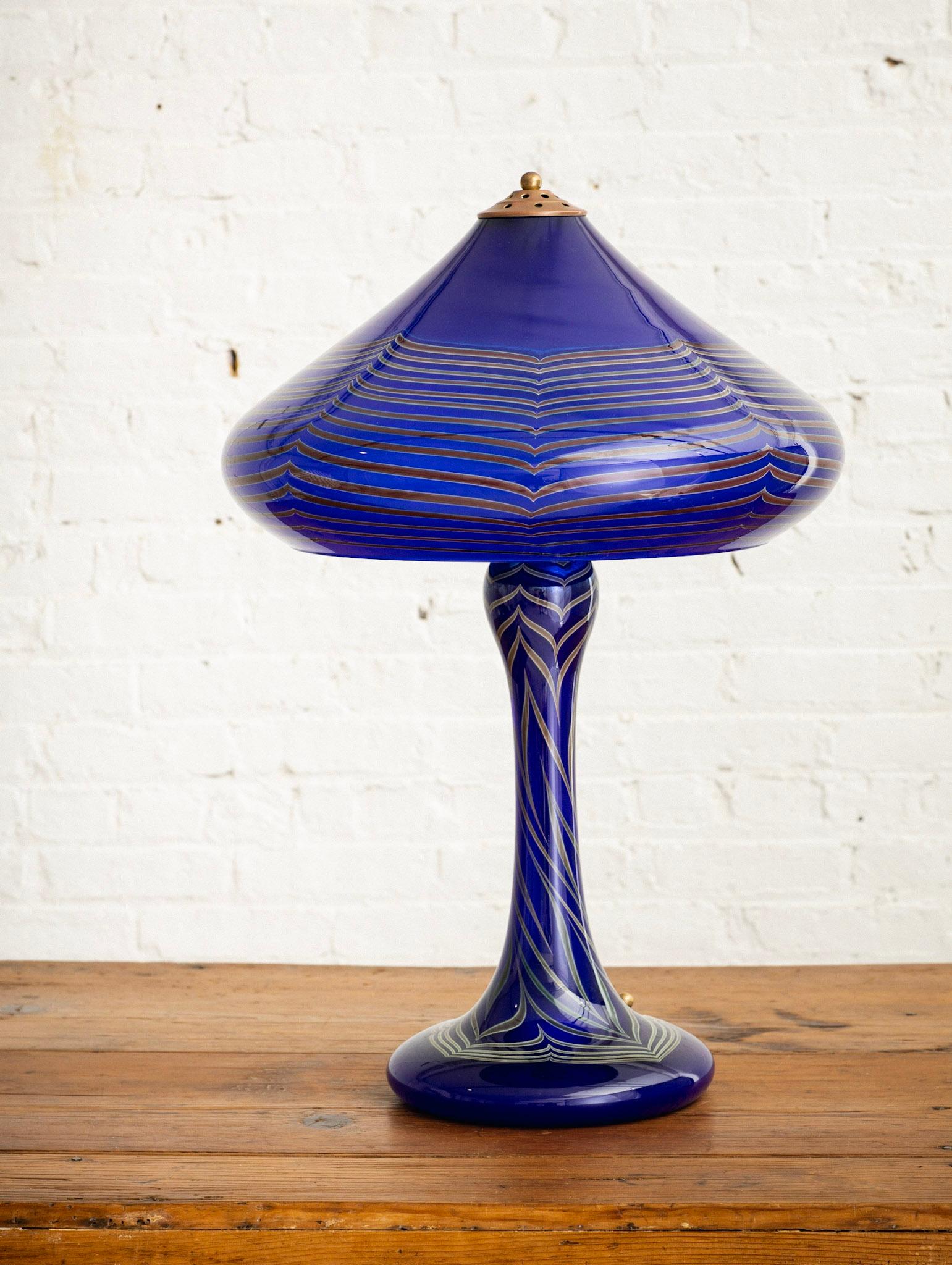 A hand blown glass table lamp by Joseph Clearman Studio. An upper glass shade is mounted to a glass bottom with brass hardware. Cobalt blue glass with a marbleized stripe detail. Art nouveau style silhouette. Upper shade is illuminated by three
