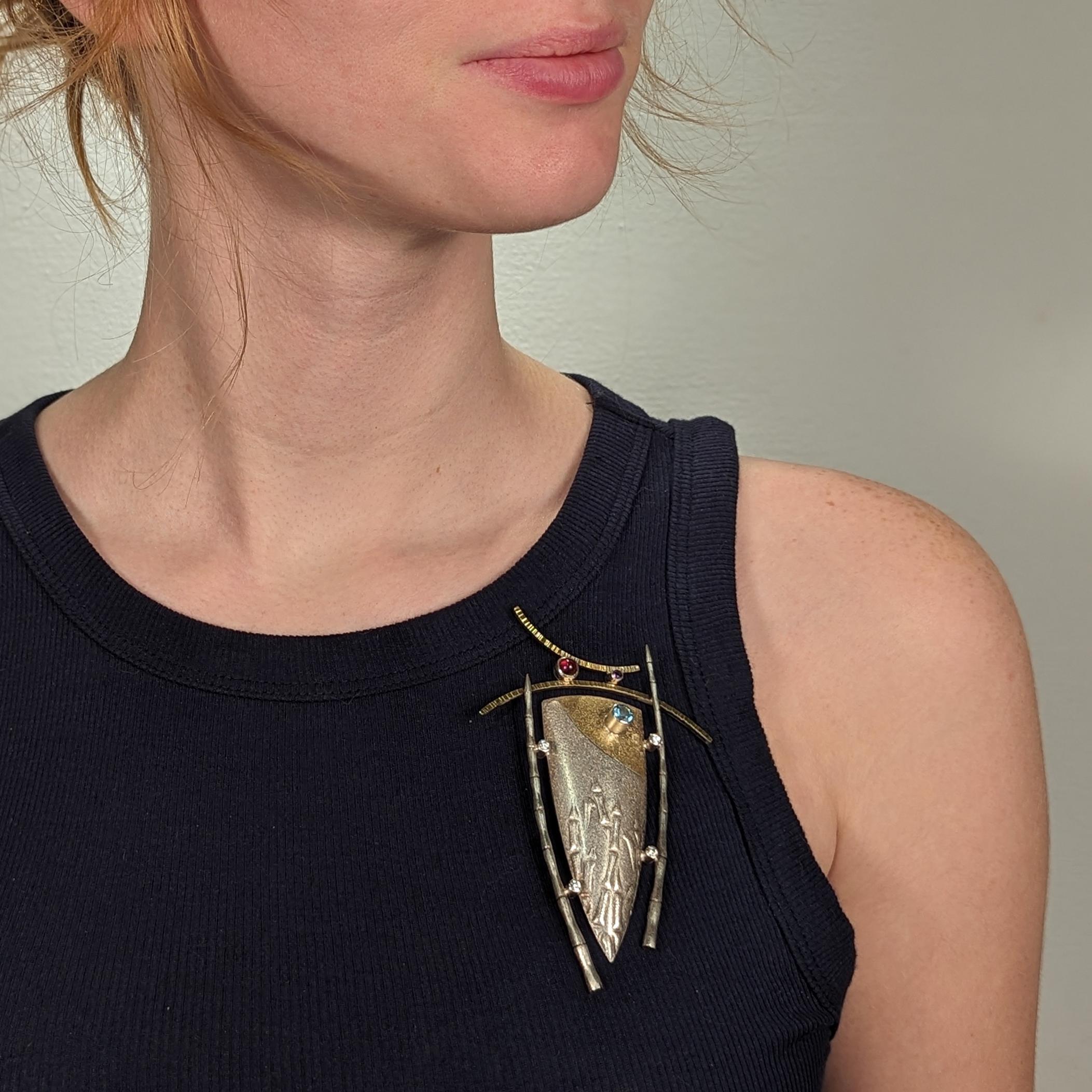 A fine Modernist gilt silver & multi-gemstone brooch.

With a parcel-gilt central shield device framed by bamboo sides, a curved top, and bezel-set with stones.

The gemstones include blue topaz, amethyst, and paste (or glass).

Marked to the