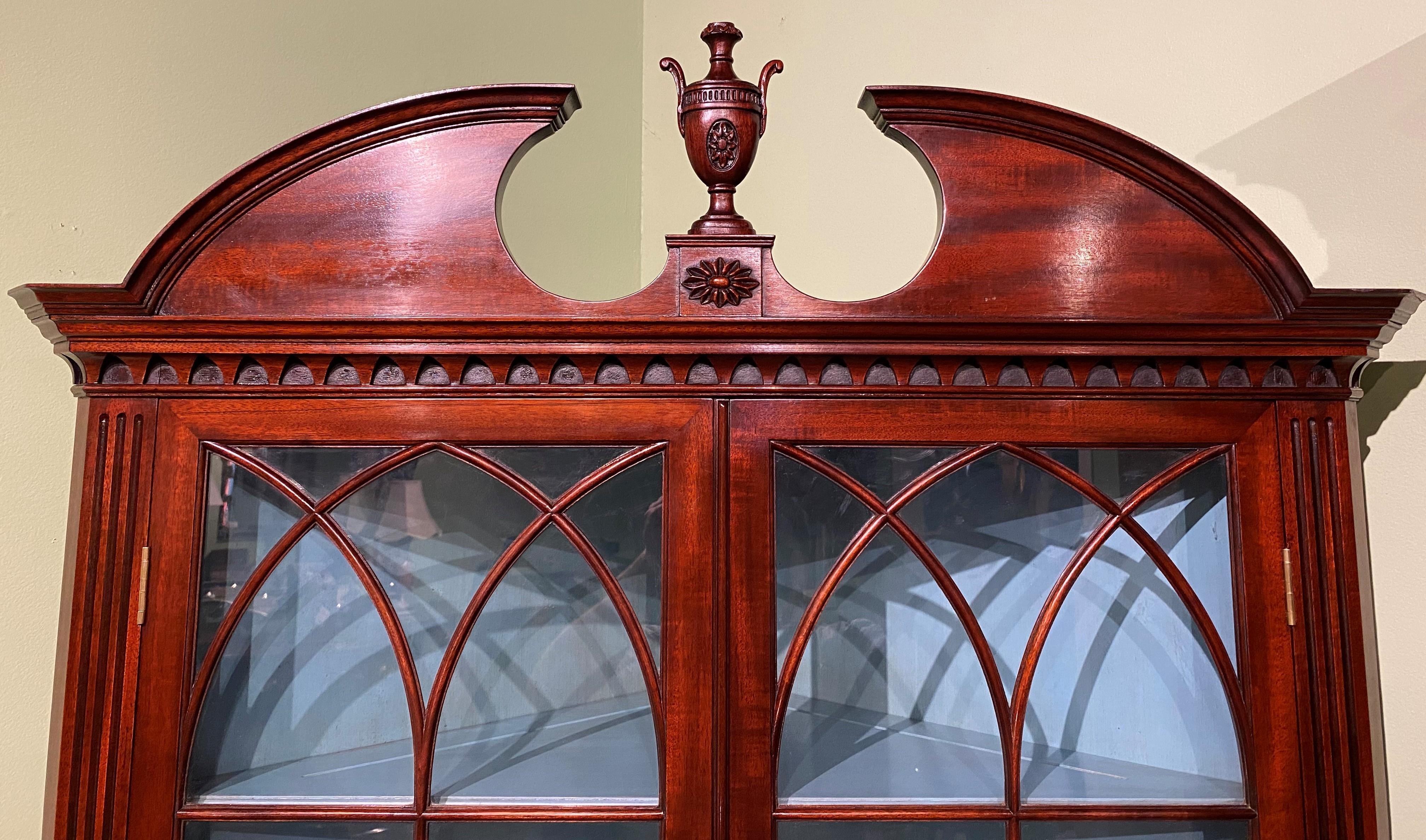 A fine quality custom mahogany one piece corner cabinet, its set back upper case with a broken pediment arched crest with central urn finial surmounting two glass doors opening to a light blue three shelf interior over a lower case with two curved