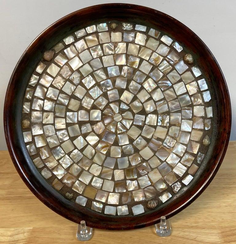Joseph Heinrich's Native American-style copper, abalone, and shell-inlaid salver
of circular form, the patinated copper body with rolled rim, and the center inset with numerous hand-cut abalone mosaic pieces in a sunburst design with seven small