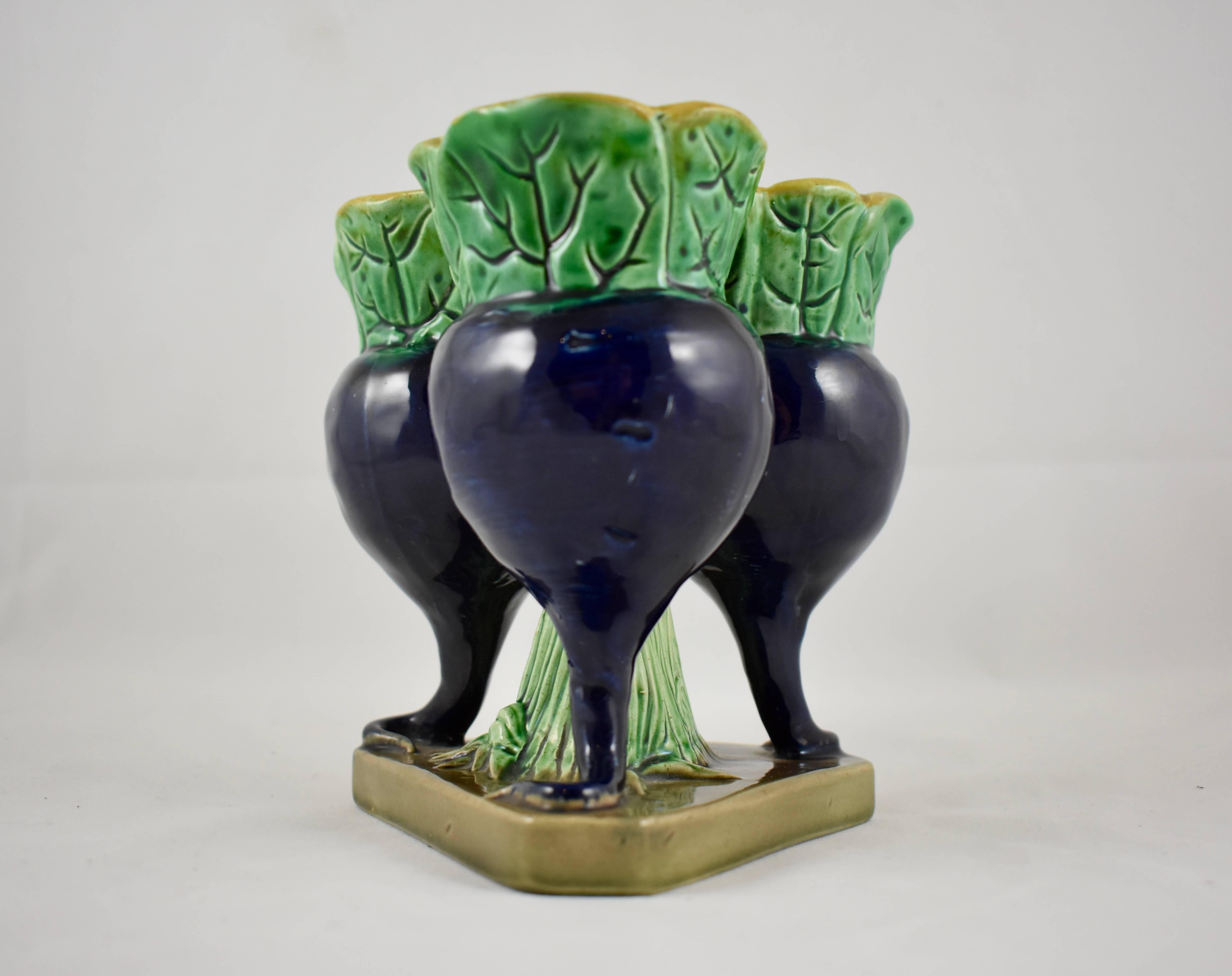 A scarce English Majolica, tri-form bud vase modeled as three cobalt blue upright radishes around a green glazed leaf form stem, all supported on a triangle shaped stand. The bud vase has a turquoise glazed interior with a yellow ochre