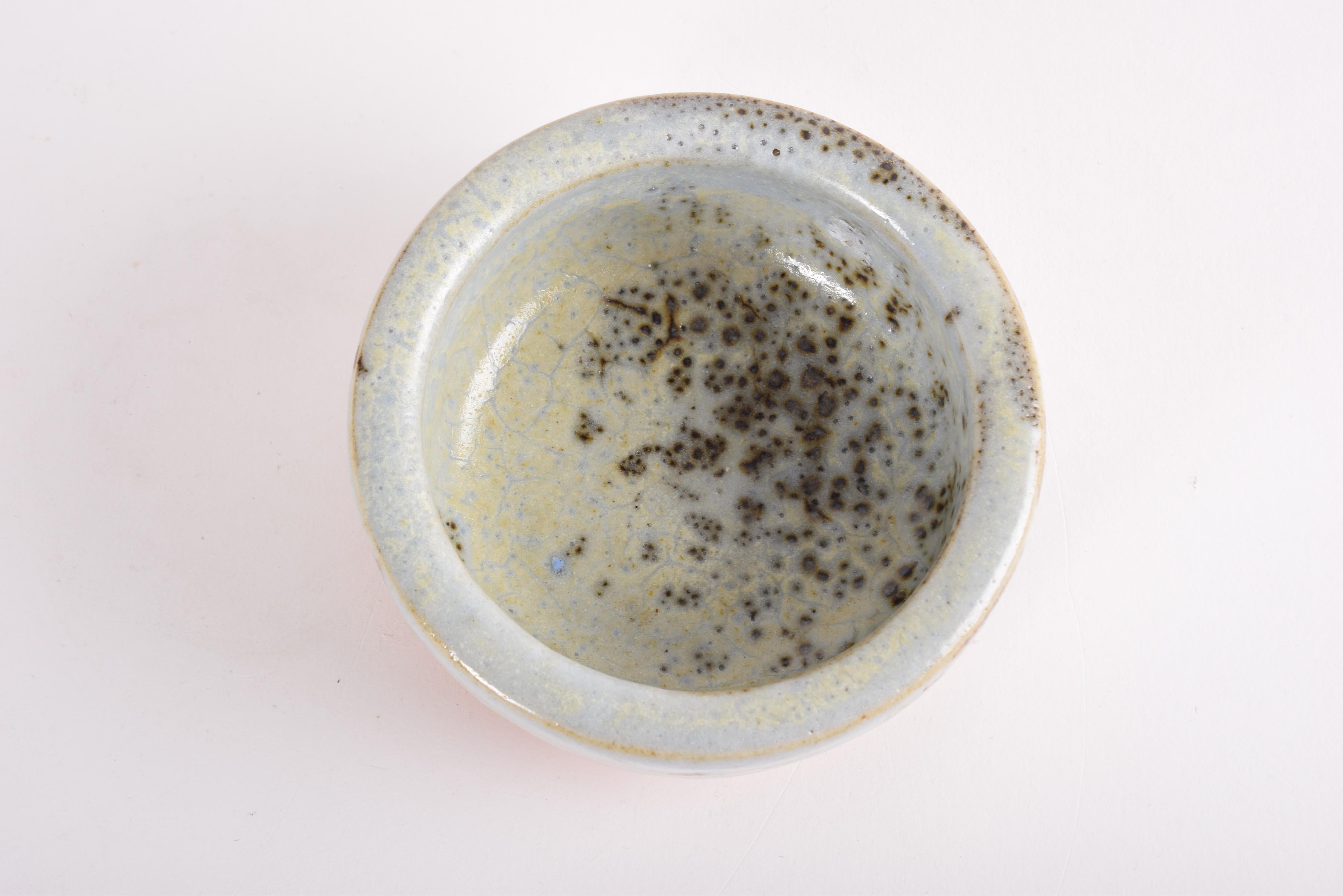 Small bowl by Danish ceramist Jørgen Mogensen (1927-2017) from his own studio. Made ca 1960s.
The bowl is made from stoneware and has thick walls. It is covered with a gray and pale blue glaze with pale yellow elements and small dark brown