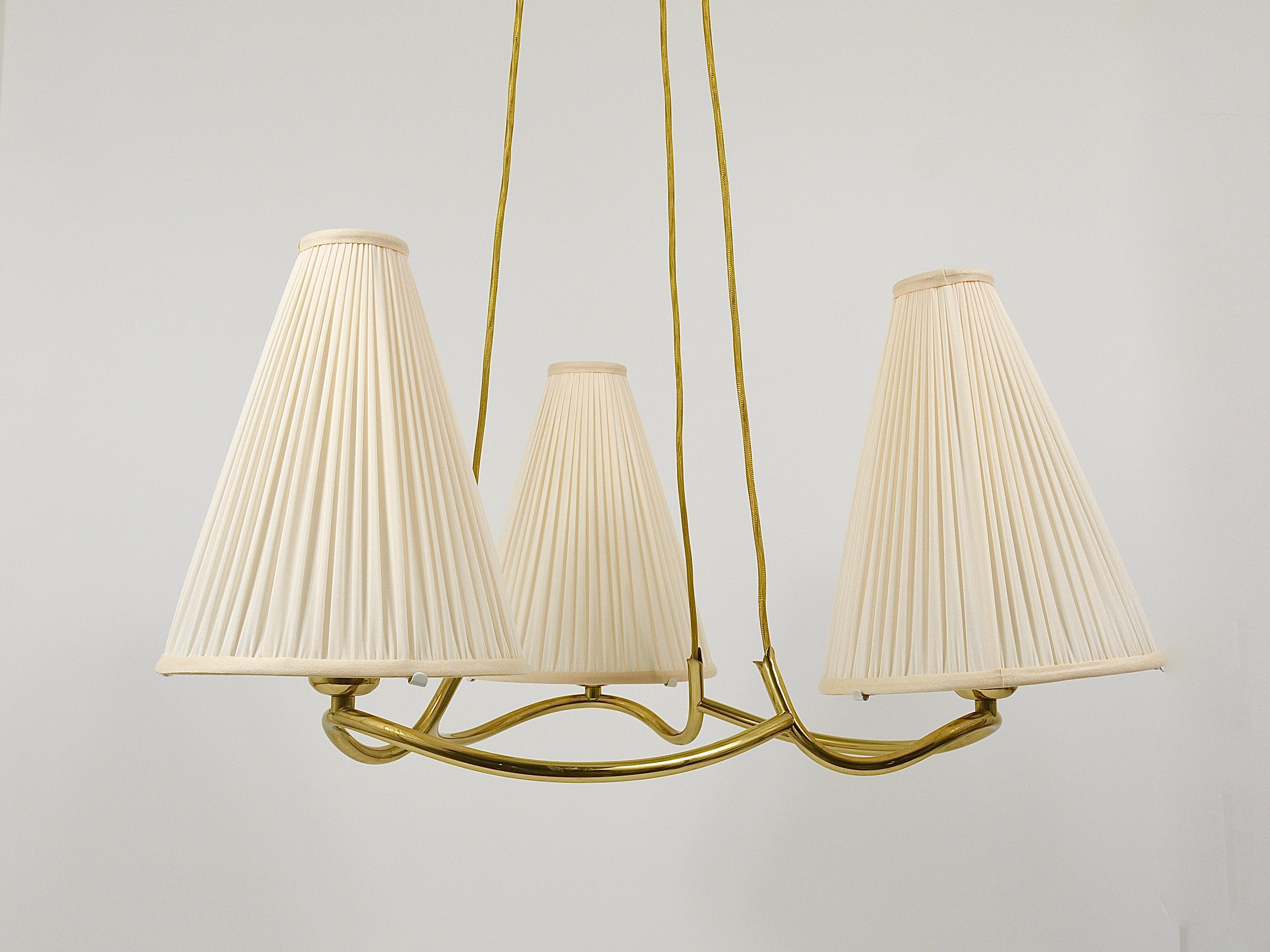 A beautiful and elegant brass chandelier, model „Heron“, manufactured by J.T. Kalmar Vienna in the early 1950s. This sculptural chandelier has a wonderfully curved brass frame, it offers three light sources with conical lampshades and hangs on