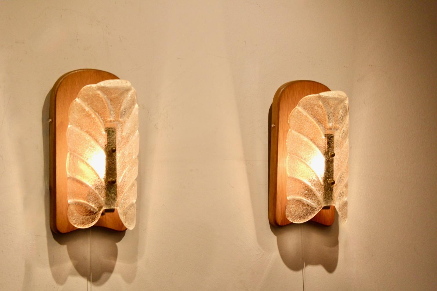 An elegant pair of wall sconces designed by J.T. Kalmar for Kalmar Werkstätten, Austria, circa 1960s. These sculptural fixtures feature thick, molded glass shades with a leaf-like motif, mounted on curved wooden backplates and accented with brass