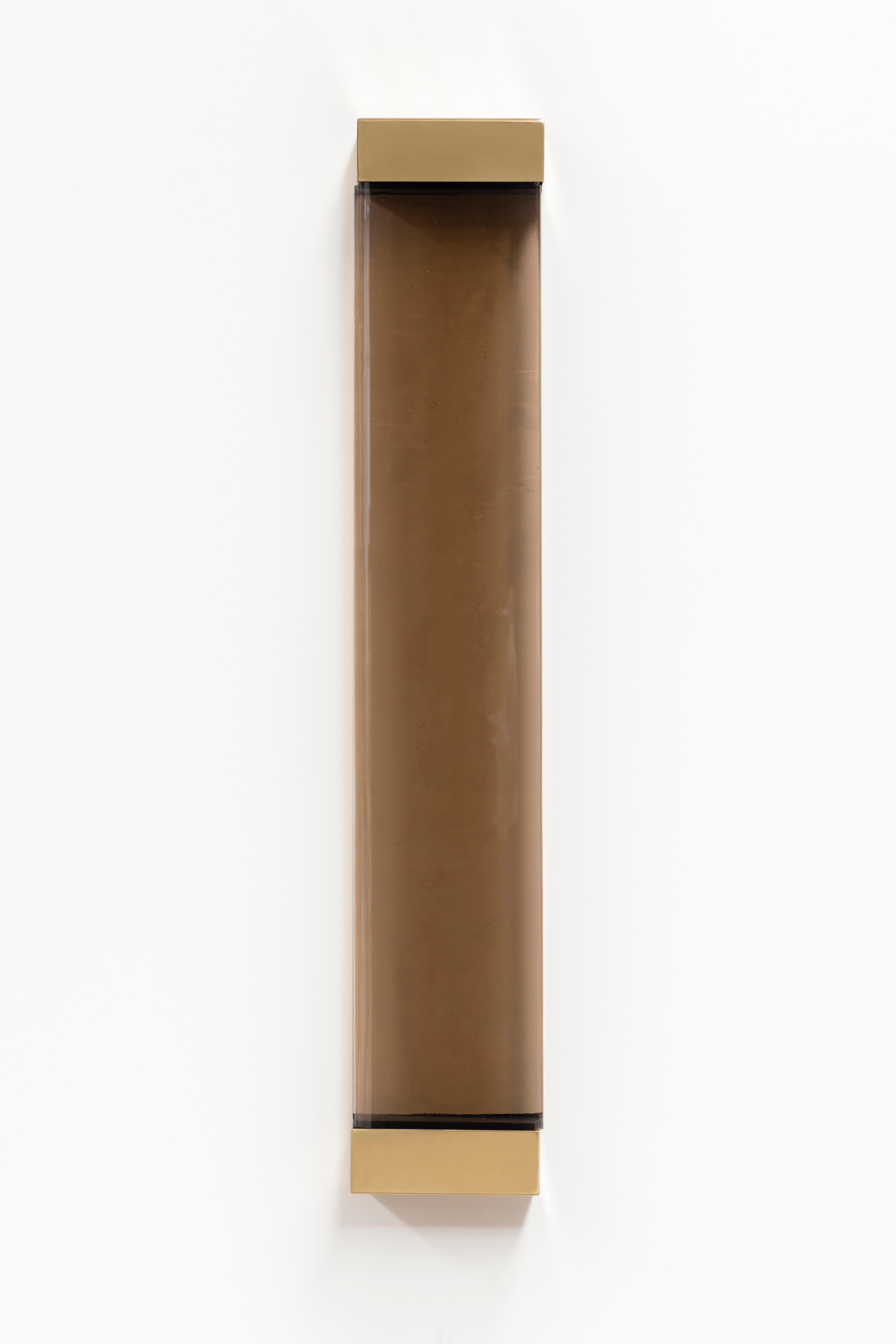 With its rigorous shapes and minimalistic contours, Jud wall lamp honors the aesthetic of Donald Judd. A geometric brass frame holds a translucent shaded glass pane concealing two colored resin filters, whose interaction with the strip LED generates