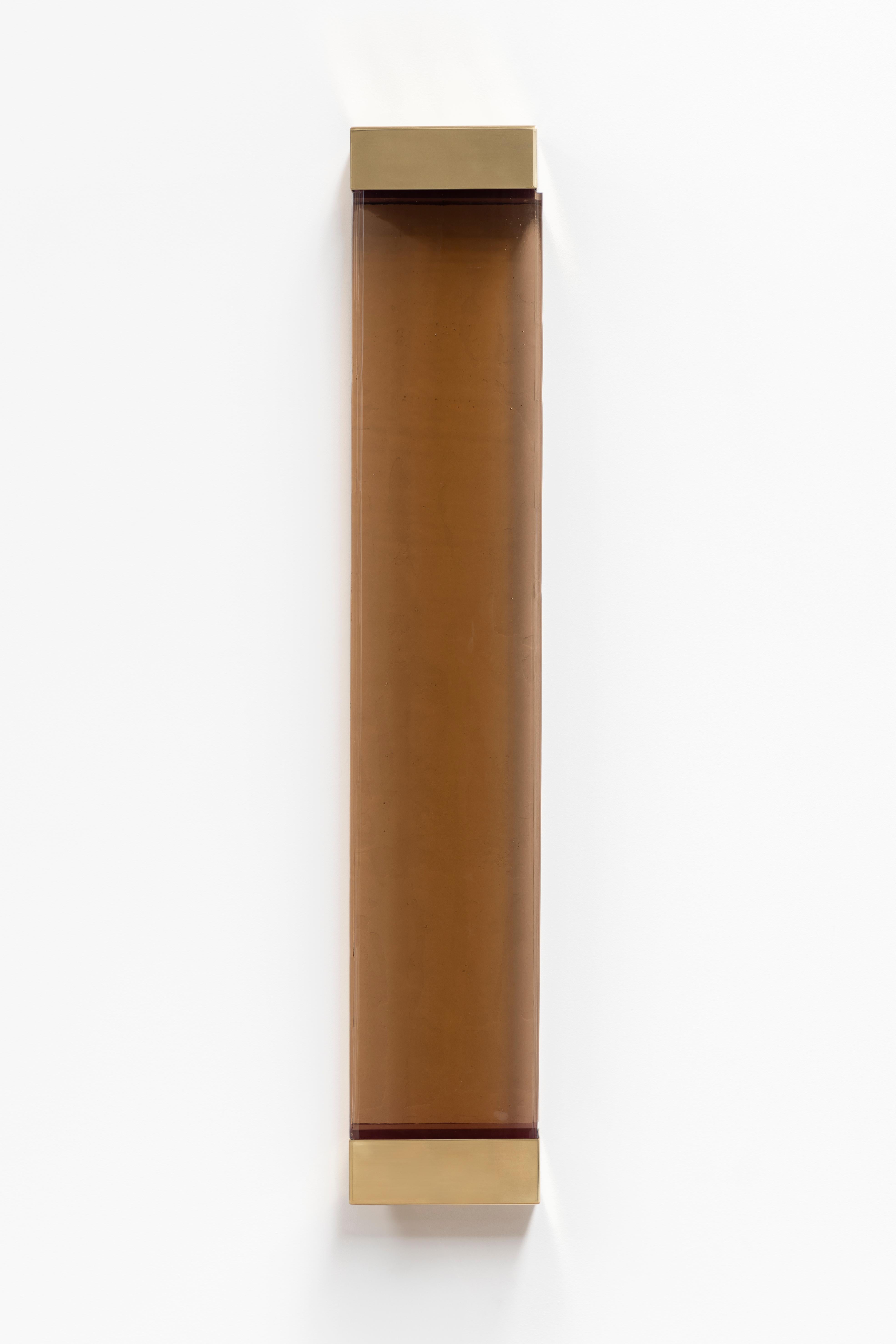 With its rigorous shapes and minimalistic contours, Jud wall lamp honors the aesthetic of Donald Judd. A geometric brass frame holds a translucent shaded glass pane concealing two colored resin filters, whose interaction with the strip LED generates