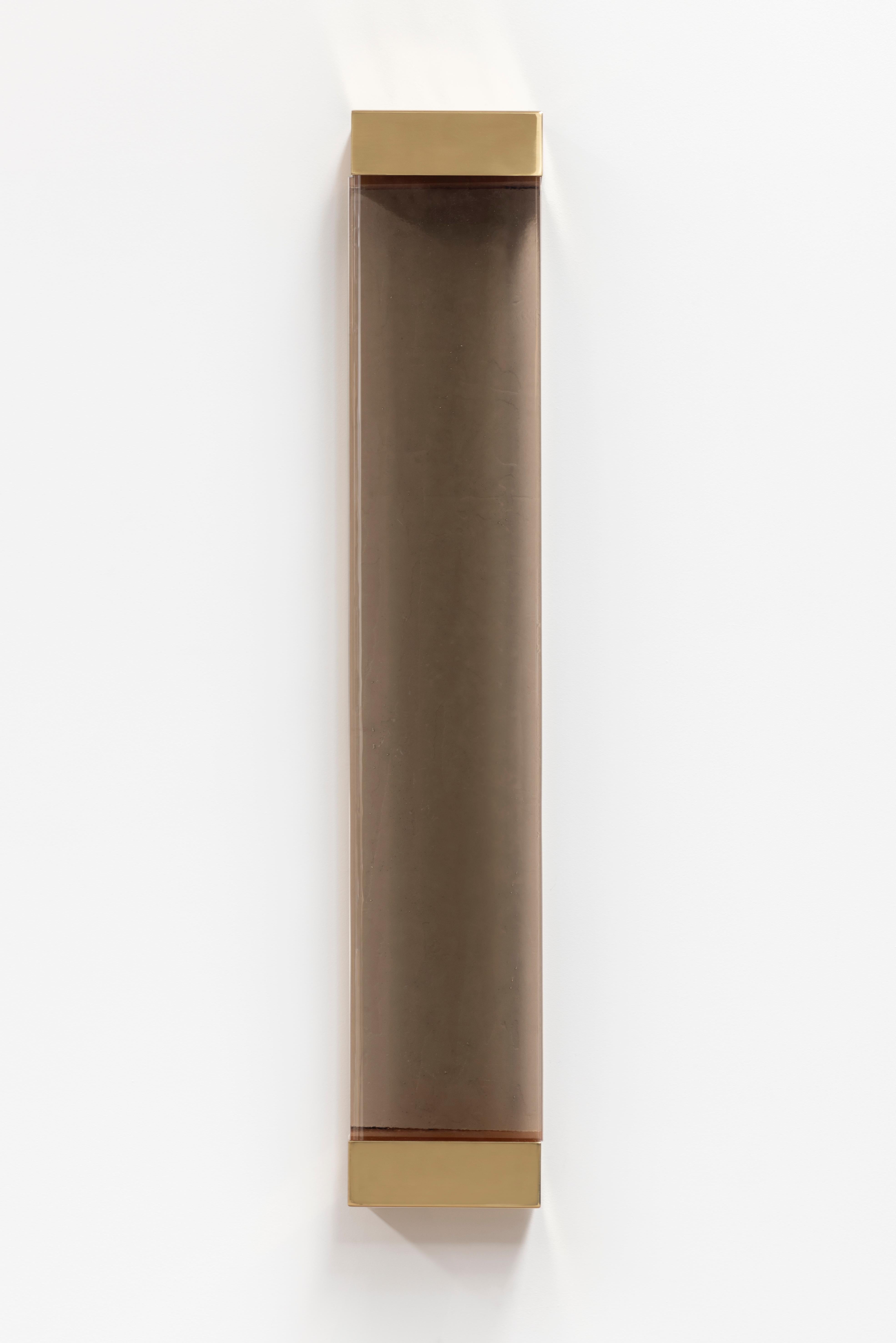 With its rigorous shapes and minimalistic contours, Jud wall lamp honors the aesthetic of Donald Judd. A geometric brass frame holds a translucent shaded glass pane concealing two colored resin filters, whose interaction with the strip LED generates