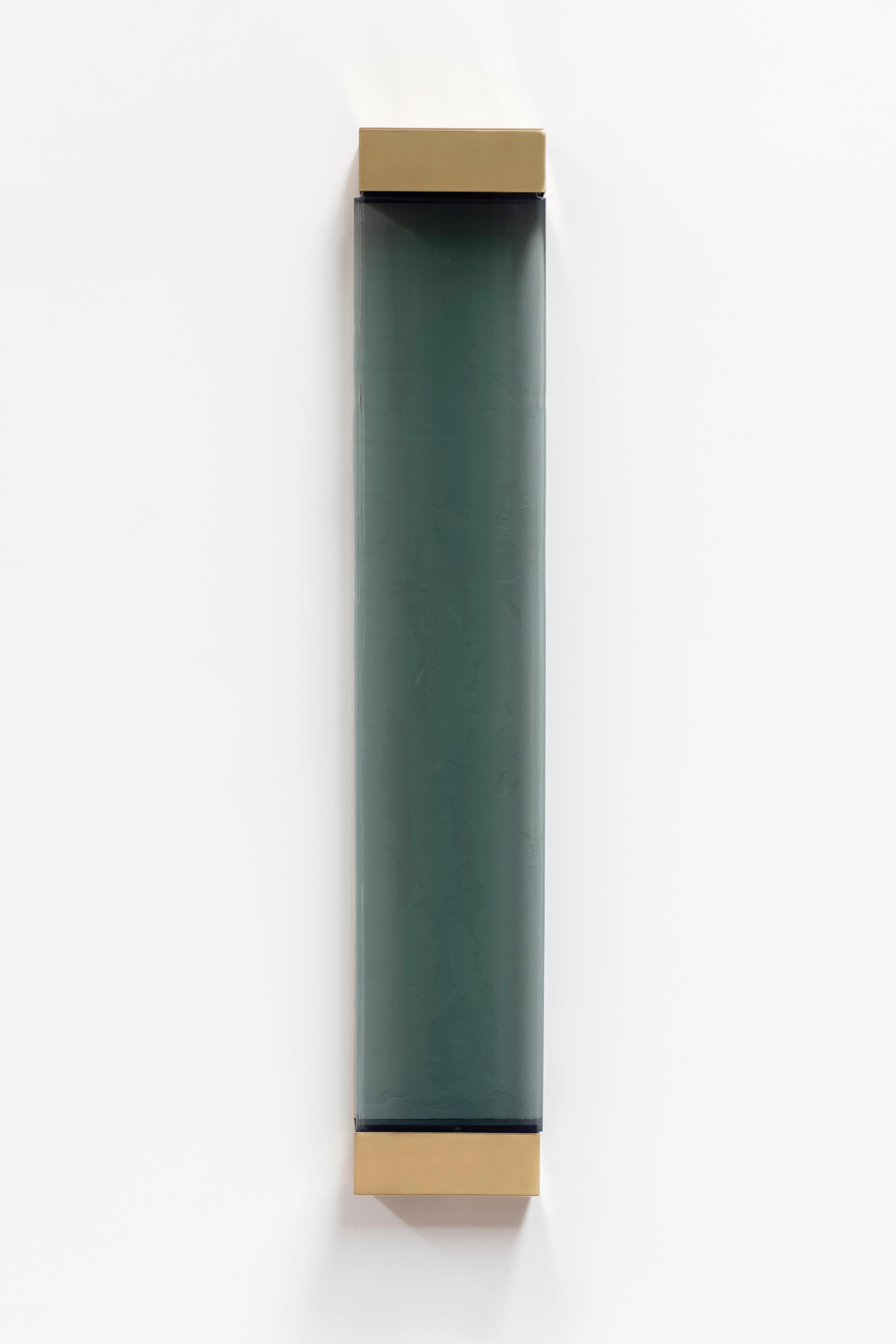 With its rigorous shapes and minimalistic contours, Jud wall lamp honors the aesthetic of Donald Judd. A geometric brass frame holds a translucent shaded glass pane concealing two colored resin filters, whose interaction with the strip LED generates