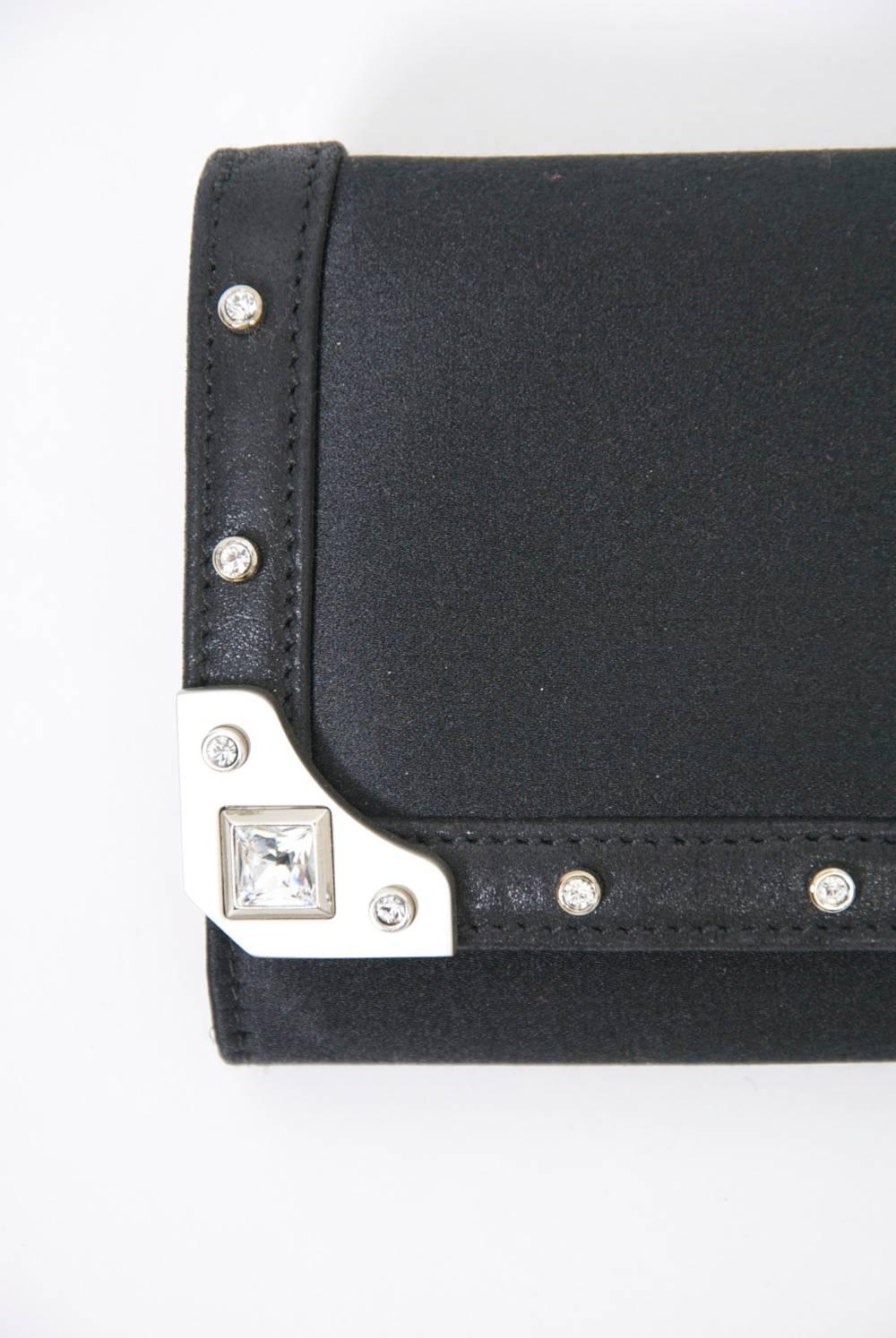Sleek rectangular black fabric clutch by Judith Leiber, the border ornamented with brushed silver metal and crystals. Magnetic clasp. Interior features attached mirror and side compartment with change purse. Dust cover.