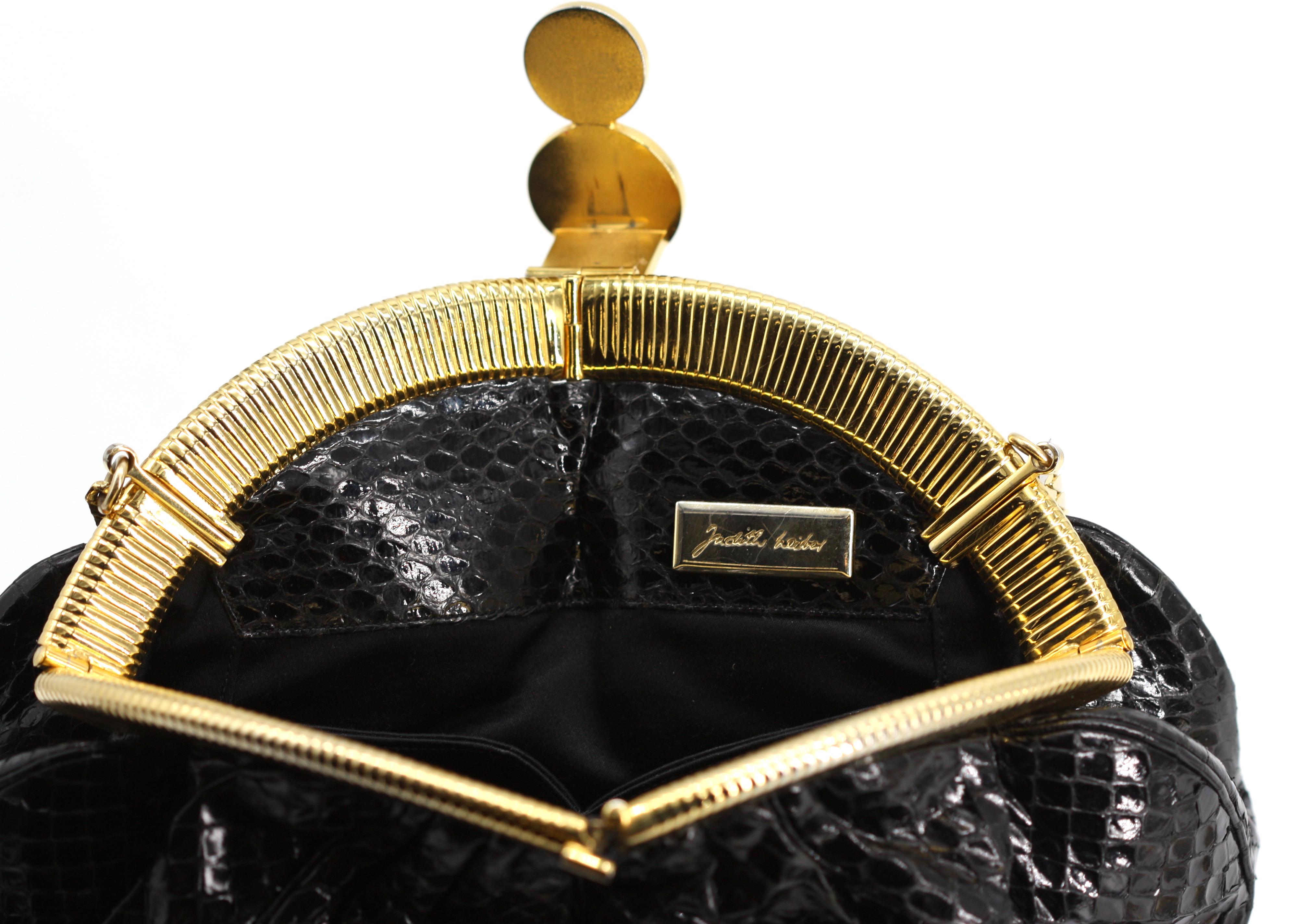
Judith Leiber Bolso negro de piel de serpiente
El bolso, en forma de saquito, tiene un borde dorado, con un cierre de esmalte rojo y negro, y una cadena de metal dorado; el interior, de piel de serpiente y tela negra, tiene una cremallera y un