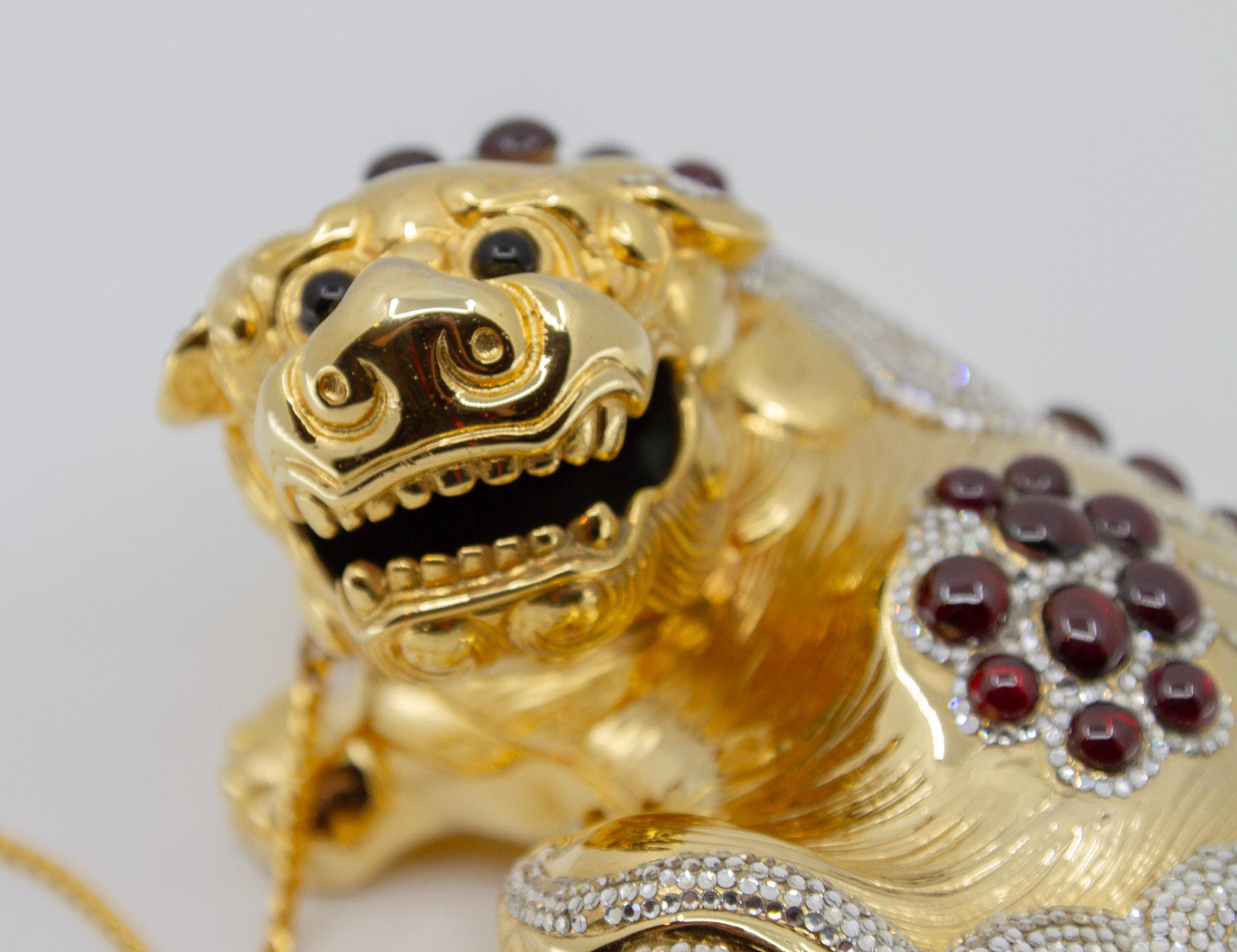 Judith Leiber Couture, United States. Chinese Ming foo dog gold-tone metal minaudiere clutch bag. With applied Swarovski crystals and cabochon garnets. The interior with gold leather, label, and gold-tone metal shoulder strap. 

Comes with miniature