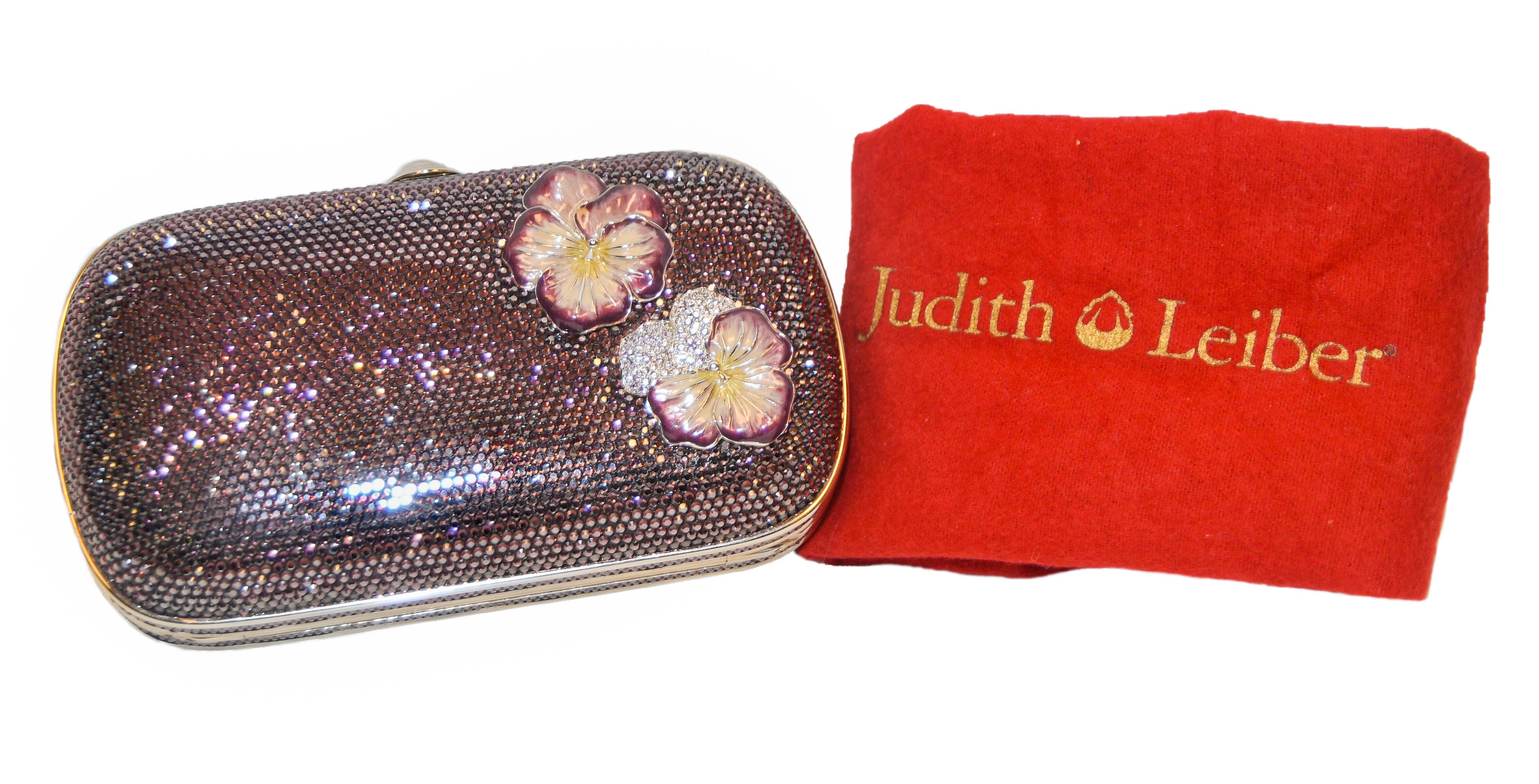 Judith Leiber purple crystal minaudiere evening clutch enhanced with 2 unique enamel Pansy flowers at front corner.  Purple Swarovski crystal exterior trimmed with silver hardware and a top push button closure. Silver leather lined interior that