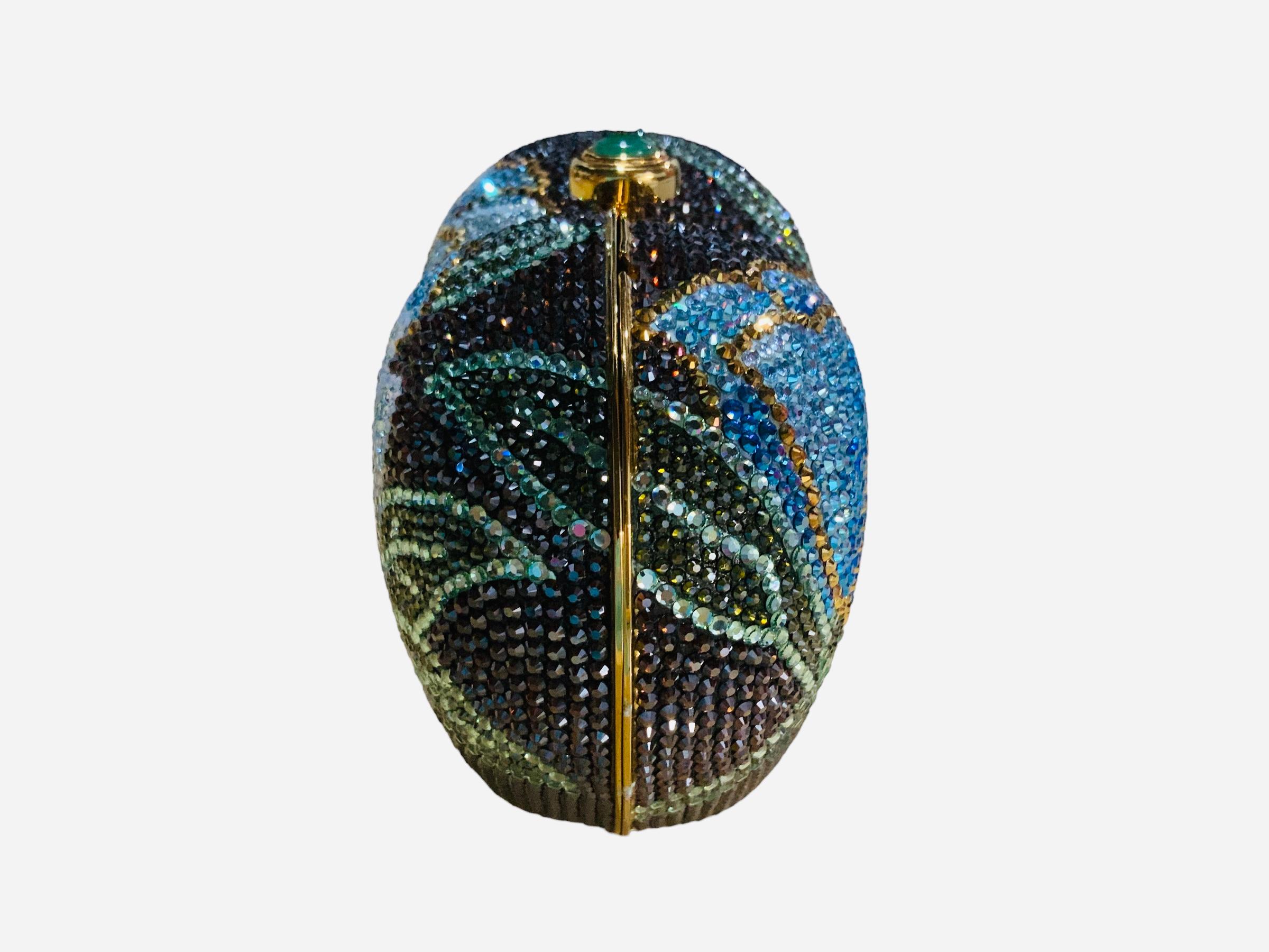 This is a Judith Leiber Swarovski crystals minaudiere / clutch. It depicts a gold tone metal heart shaped minaudiere adorned with a large peony flower with green leaves in the front and a semi close one in the back. Long branches of green leaves