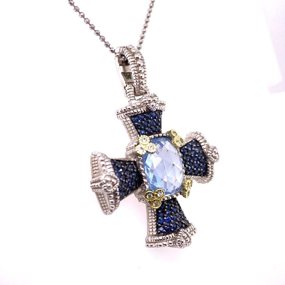 Colorful Cross Pendant by designer Judith Ripka. The pendant is fashioned in sterling silver and 18 karat yellow gold. The center features a faceted blue topaz gemstone, and the pendant measures 1.25 x 1.25 inches (1.75 inches with the baile).