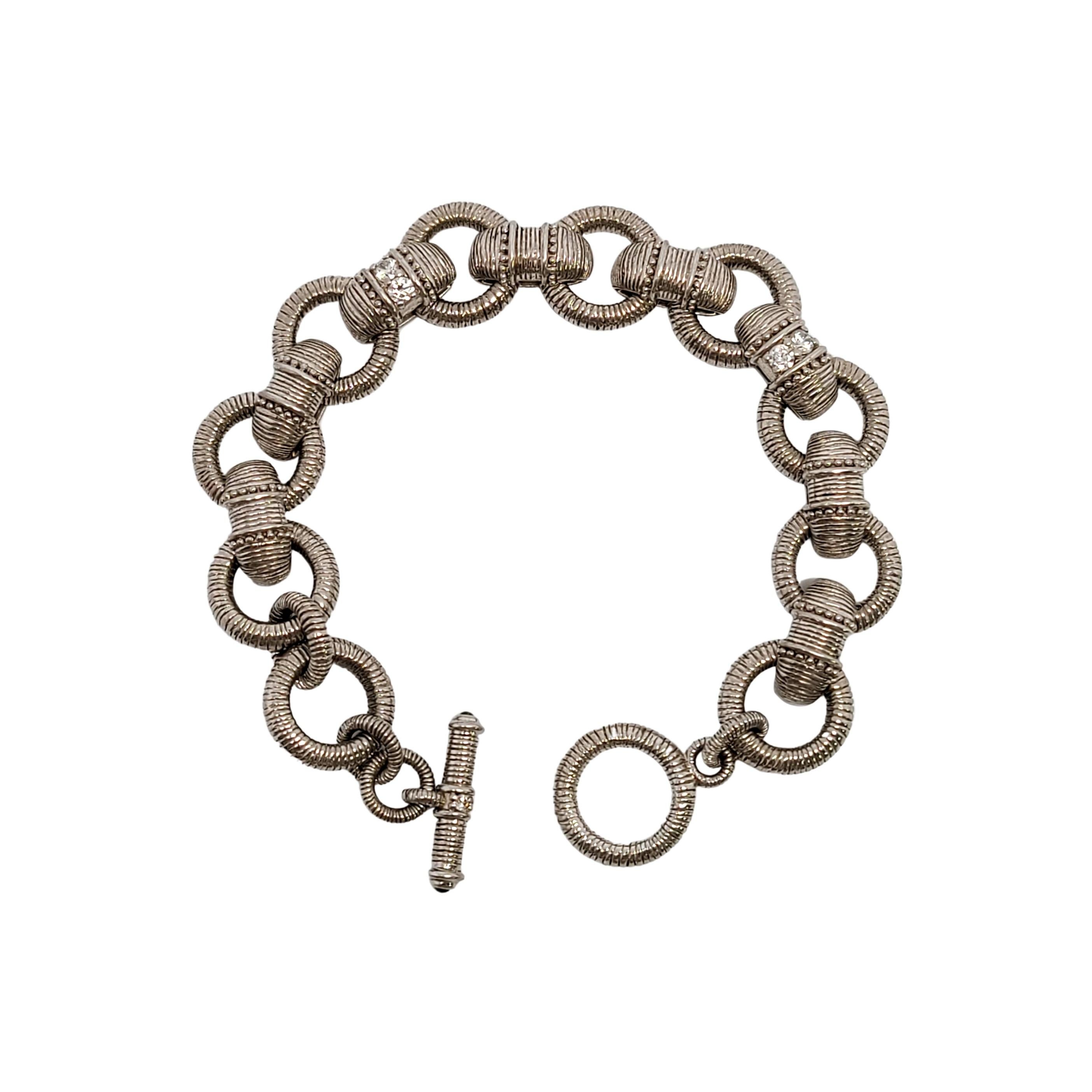 Sterling silver textured link and CZ Diamonique toggle bracelet by designer Judith Ripka.

This beautiful piece features circle and elongated textured links, with a toggle closure that has Diamonique CZ accents and a small green stone on each end of