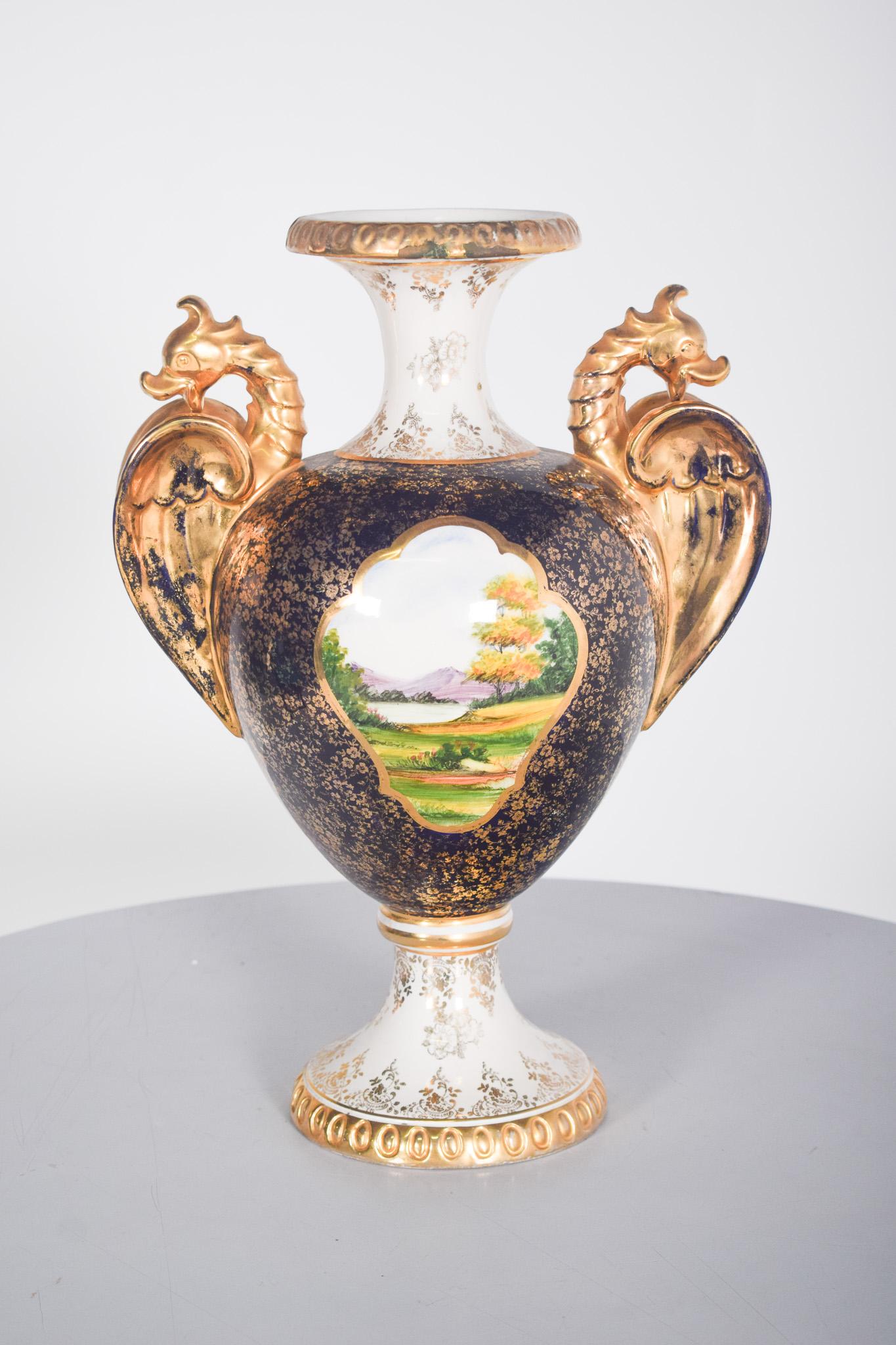 Brocca con manici in ceramica portoghese di Alcobaça, dipinta a mano. Decorazione policroma e dorata, con un paesaggio al centro all'interno di un medaglione, piccoli fiori dorati in tutta la decorazione.
Le ali in rilievo raffiguranti draghi alati