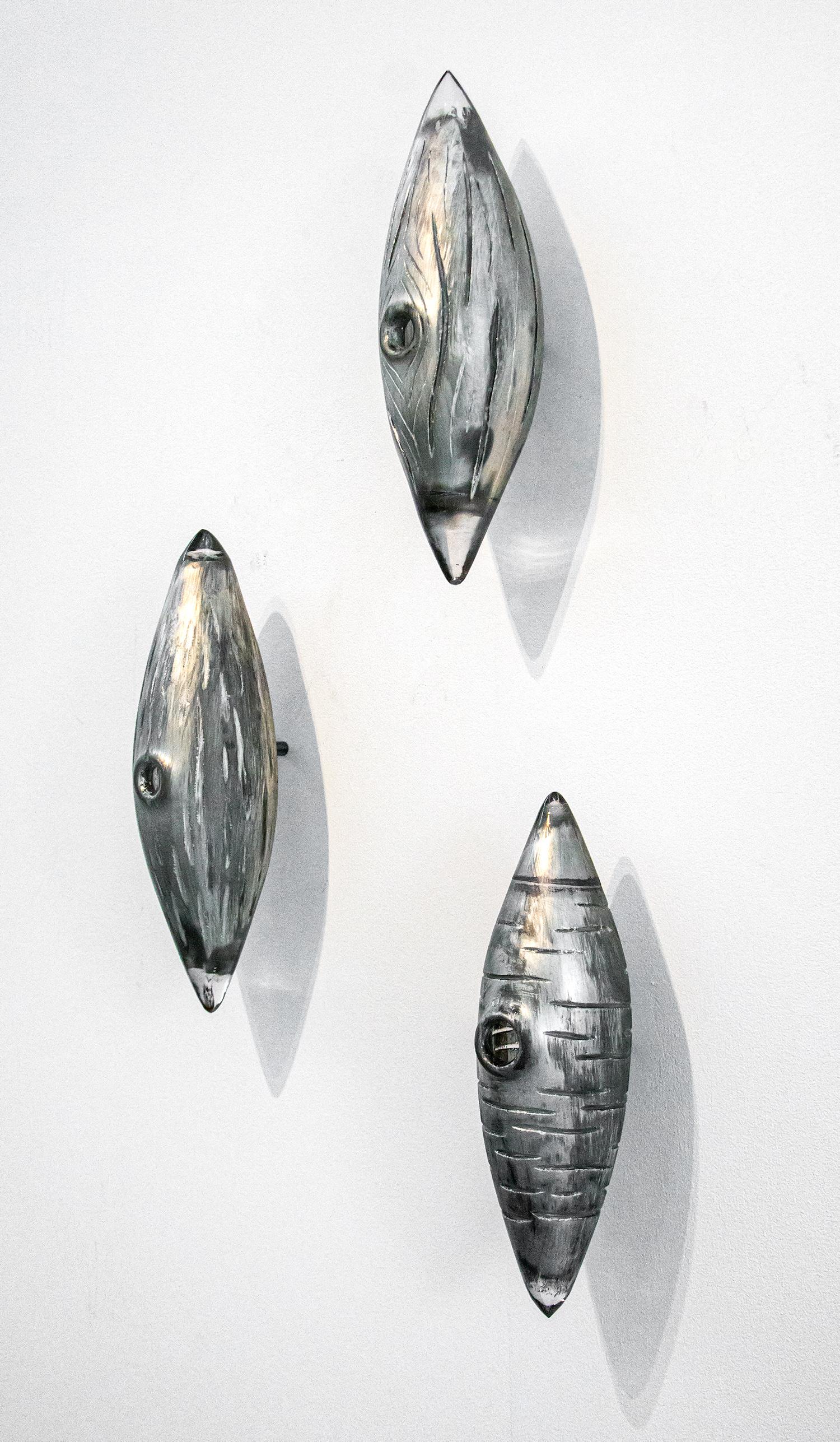 Three glass cocoon shapes in clear and silver with a woodgrain texture are curated vertically in this unique wall sculpture by Julia Reimer. Inspired by nature, the elements resemble cocoons or because they are each pierced with a round hole, bird's