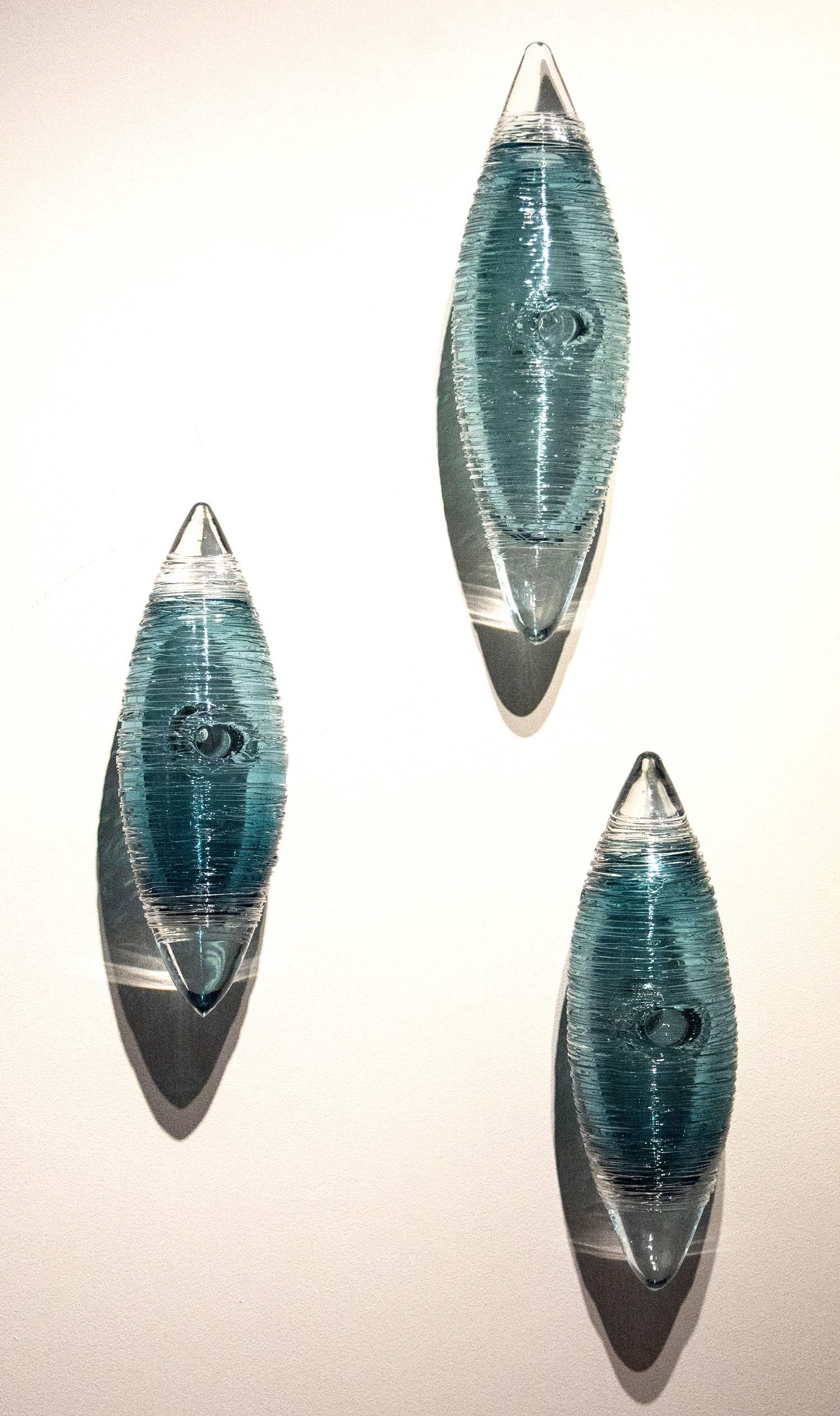 Threads of clear on translucent steel blue glass create shimmering shadows on the wall behind this grouping of three cocoons by Canadian artist Julia Reimer. Each cocoon shape features a central hole, like that of a bird's nest such as the oriole.