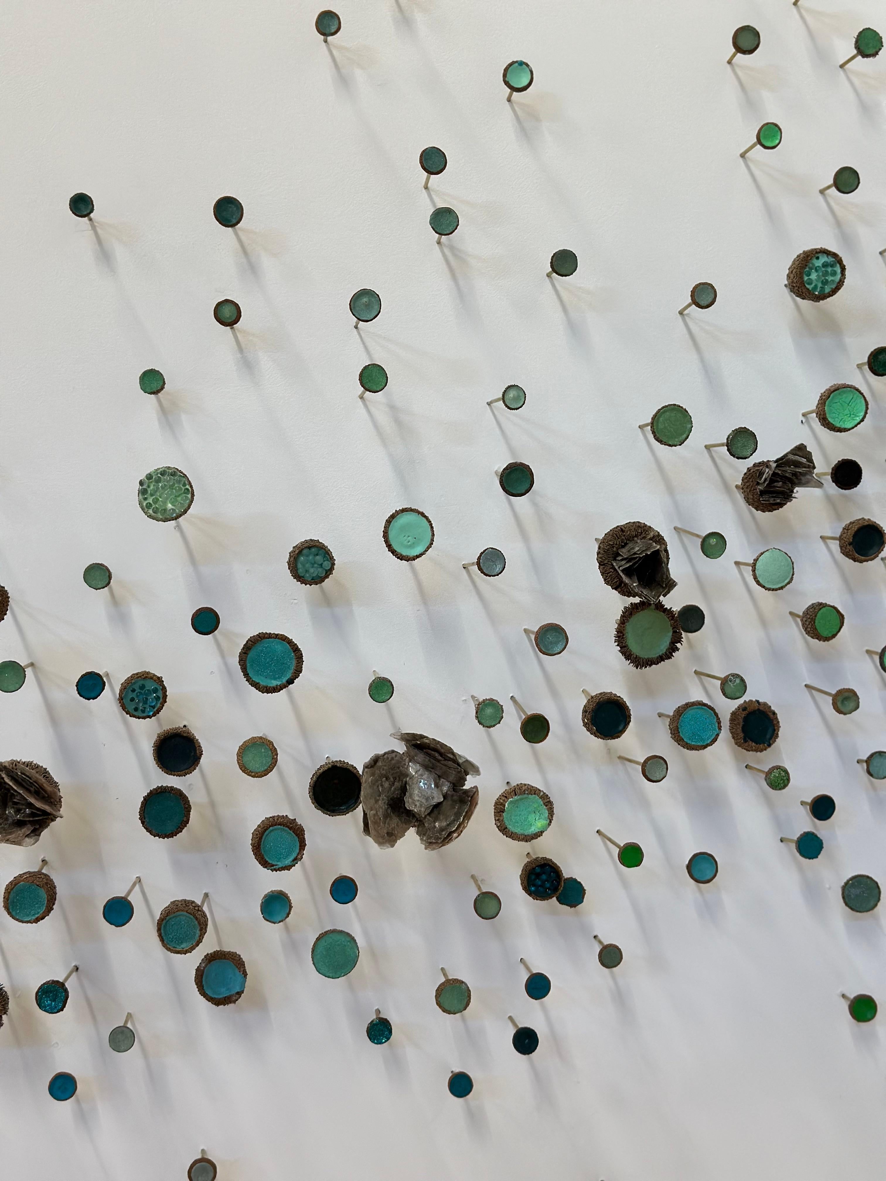 Questa installazione scultorea e serpeggiante a parete, destinata a un angolo, è composta da cappucci di ghianda su perni di ottone riempiti di vernice acrilica, cristalli naturali e mica, vetro, glitter, ottone e materiali sintetici in tonalità