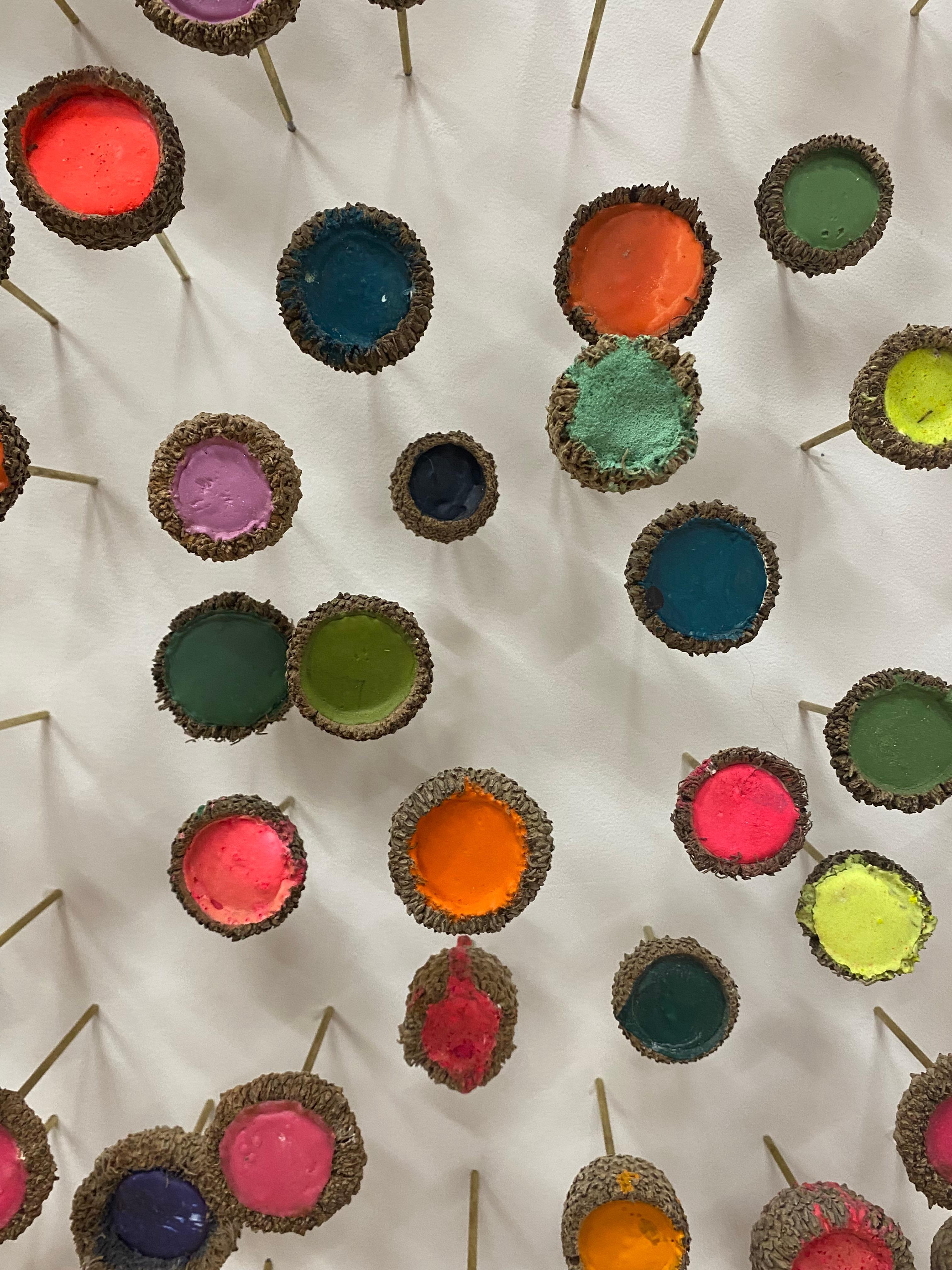 Esta instalación mural escultórica se compone de más de 275 capuchones de bellota individuales montados sobre clavijas de latón; cada capuchón está relleno de yeso y pintura en tonos brillantes de rosa, verde, morado, naranja y amarillo neón, en