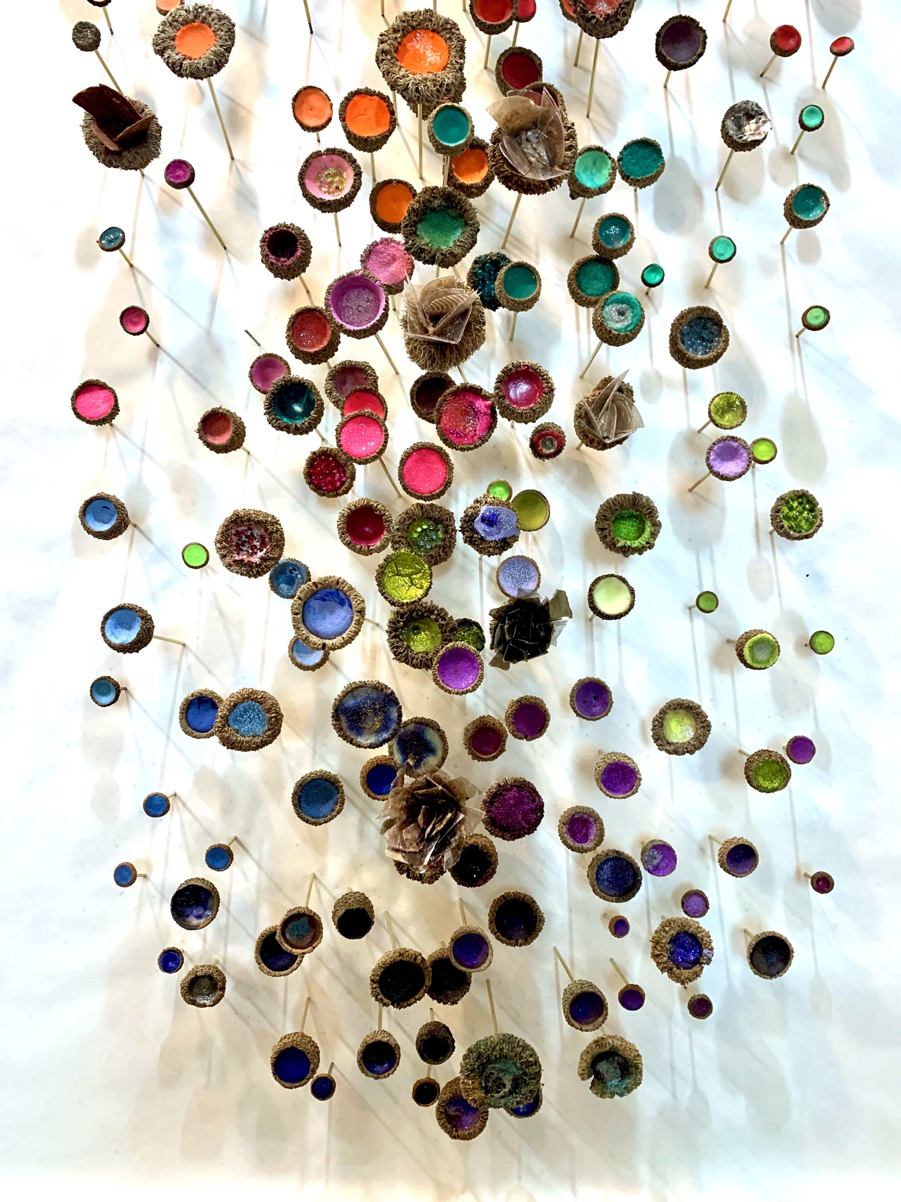 Large, vertical wall sculpture consisting of a meandering installation of acorn caps on brass pins filled with acrylic paint, natural crystals and mica, glass, glitter, brass, and synthetic materials in bright shades of luminous yellow, pink,