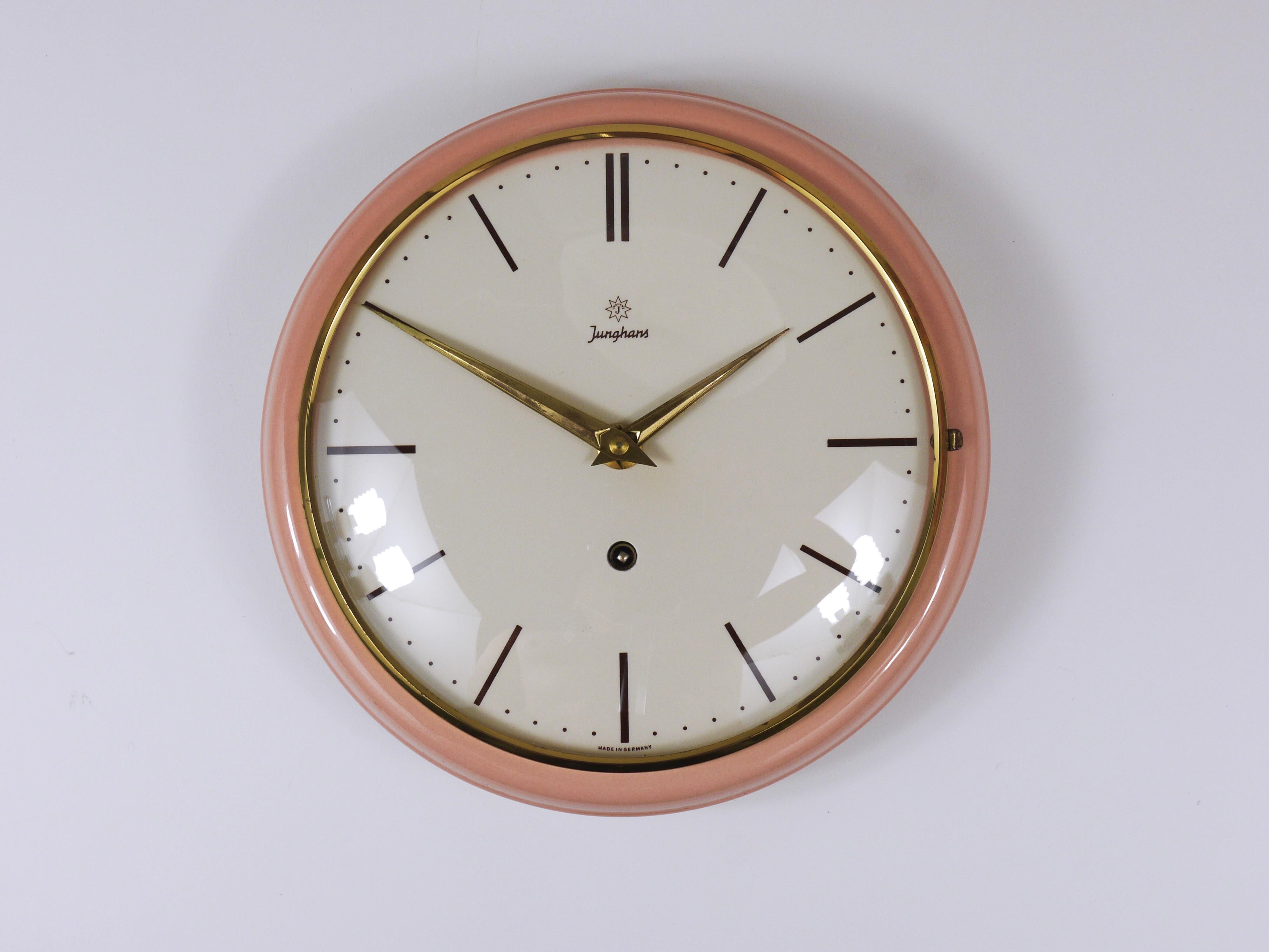 A lovely round mid-century wall clock, crafted by Junghans in Germany during the 1950s. Designed in the characteristic style of the era, it features elegant brass hands, a brass frame with domed glass, and a rose pink–melon peach ceramic housing.