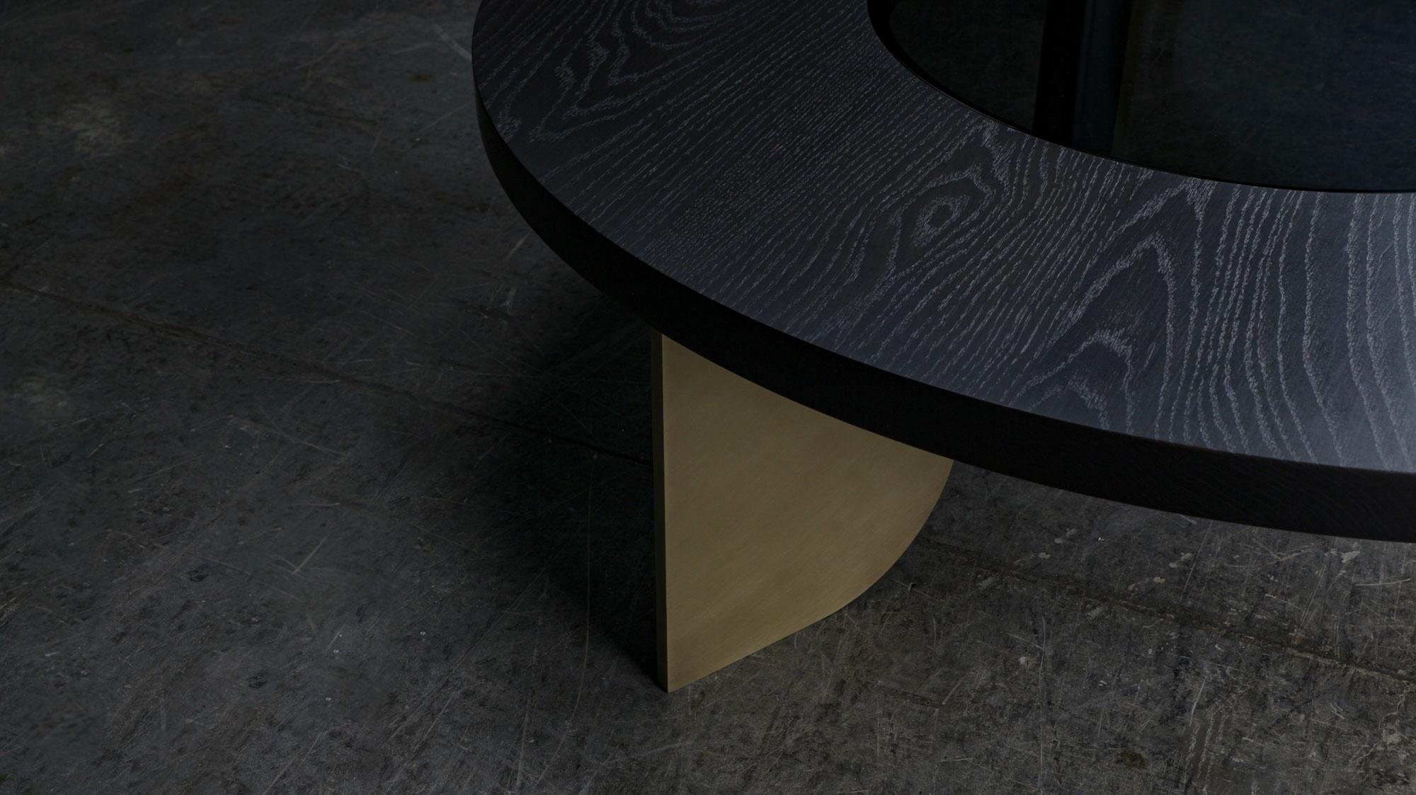 La table basse Jupiter est fabriquée à la main à partir d'une épaisse surface en bois massif et comporte une surface circulaire en verre teinté qui éclaircit la surface et permet une certaine réflexion de la lumière. La surface en bois massif est
