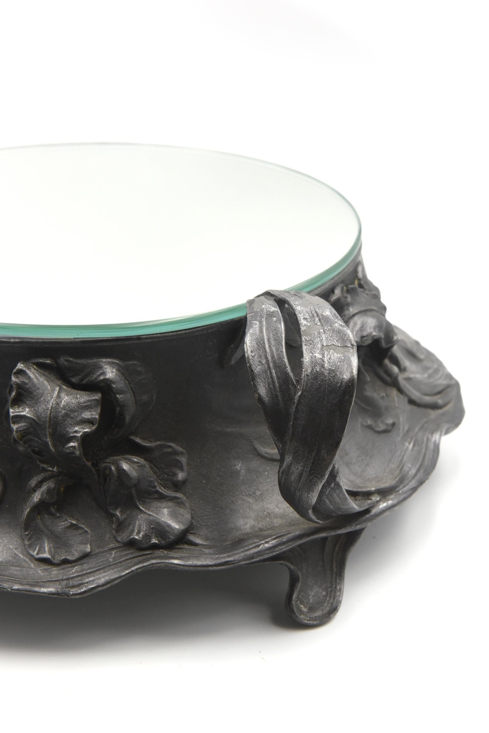 Ornate raised floral design Art Nouveau plateau or cake stand centrepiece made by Juventa in pewter with a mirror on top.  The mirrored area for the cake is 8” in diameter.

Juventa
Hans Peter started making designs for the WMF factory in 1884 from