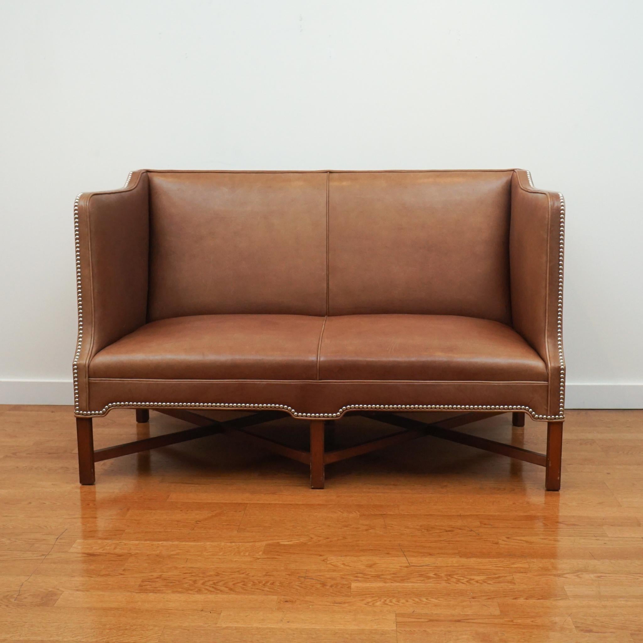 This Kaare Klint-style leather sofa is a compact, two-seater version of a design that was created for the Danish Prime Minister’s office in 1930. Featuring high side arms, deep seat and exposed, cross legged mahogany frame, the tight seat and back