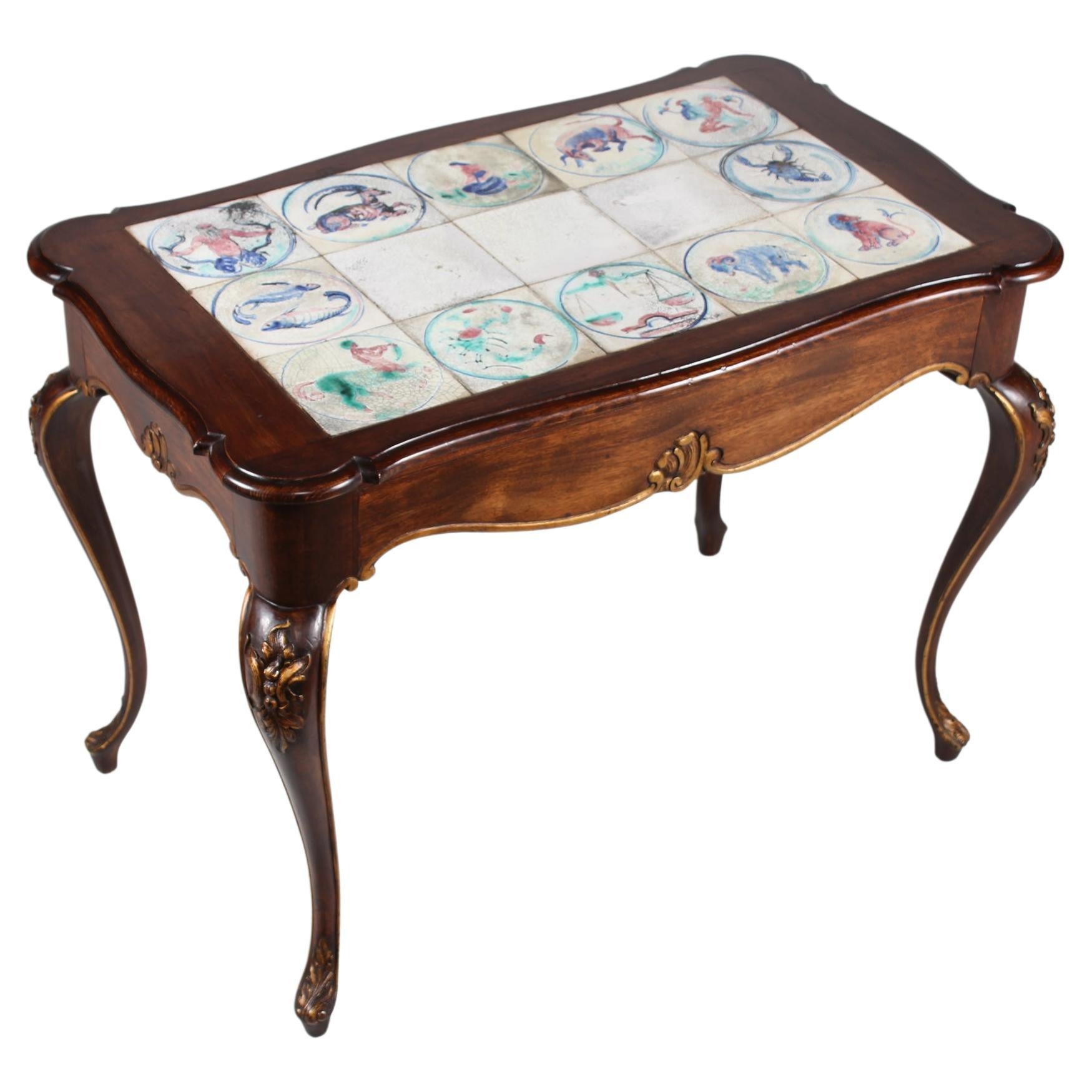 Danish Rococo-style table made of mahogany with curved legs and gilded carvings of stylized foliage. 

The tiles are decorated by Jens Thirslund (1892-1942) with the 12 zodiac signs, 1 for each month.
Jens Thirslund was for years a talented and much