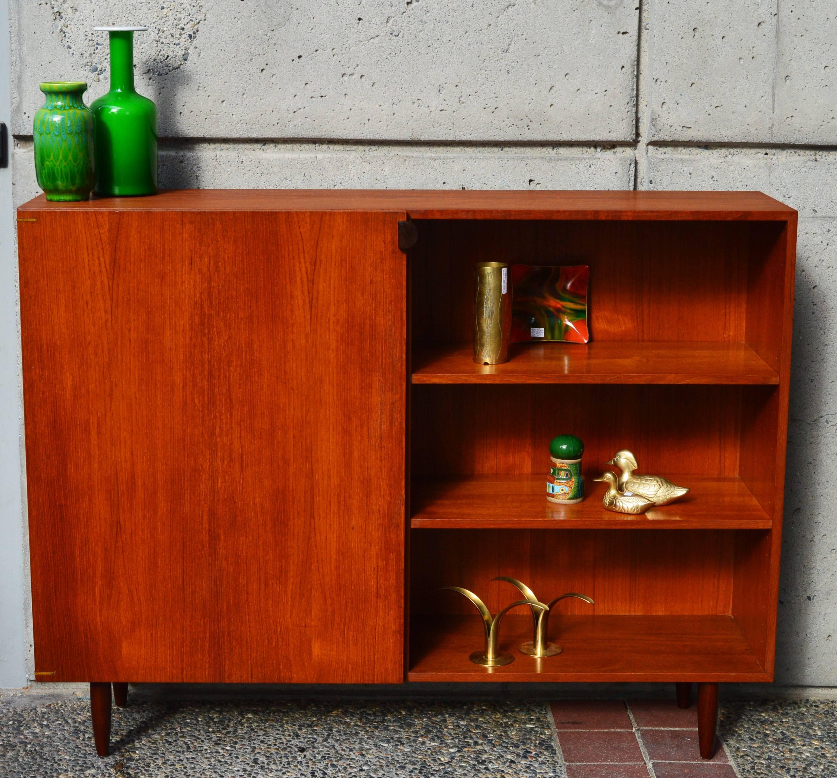 This rare sized Danish modern teak quality cabinet was designed by Kai Kristiansen for Feldballes Mobelfabrik and features a narrow profile, four adjustable shelves - with one side open and one side hidden behind a door. The door has a carved