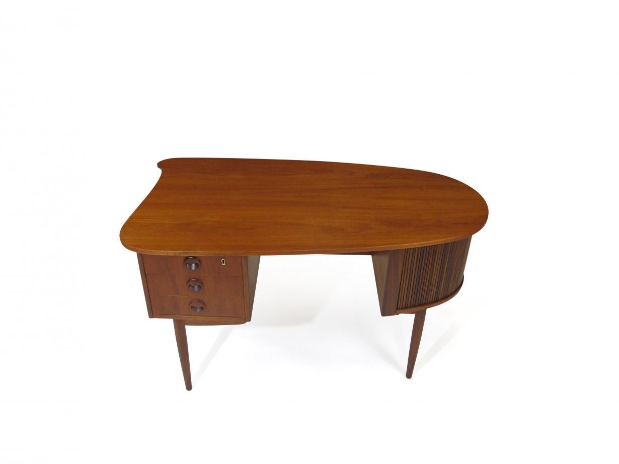Midcentury teak desk designed by Kai Kristiansen, Model 54, crafted of teak, with a bullet shaped top surface, series of three drawers with craved pulls, right side tambour door cabinet with hidden drawer and book shelf on back side of the desk,