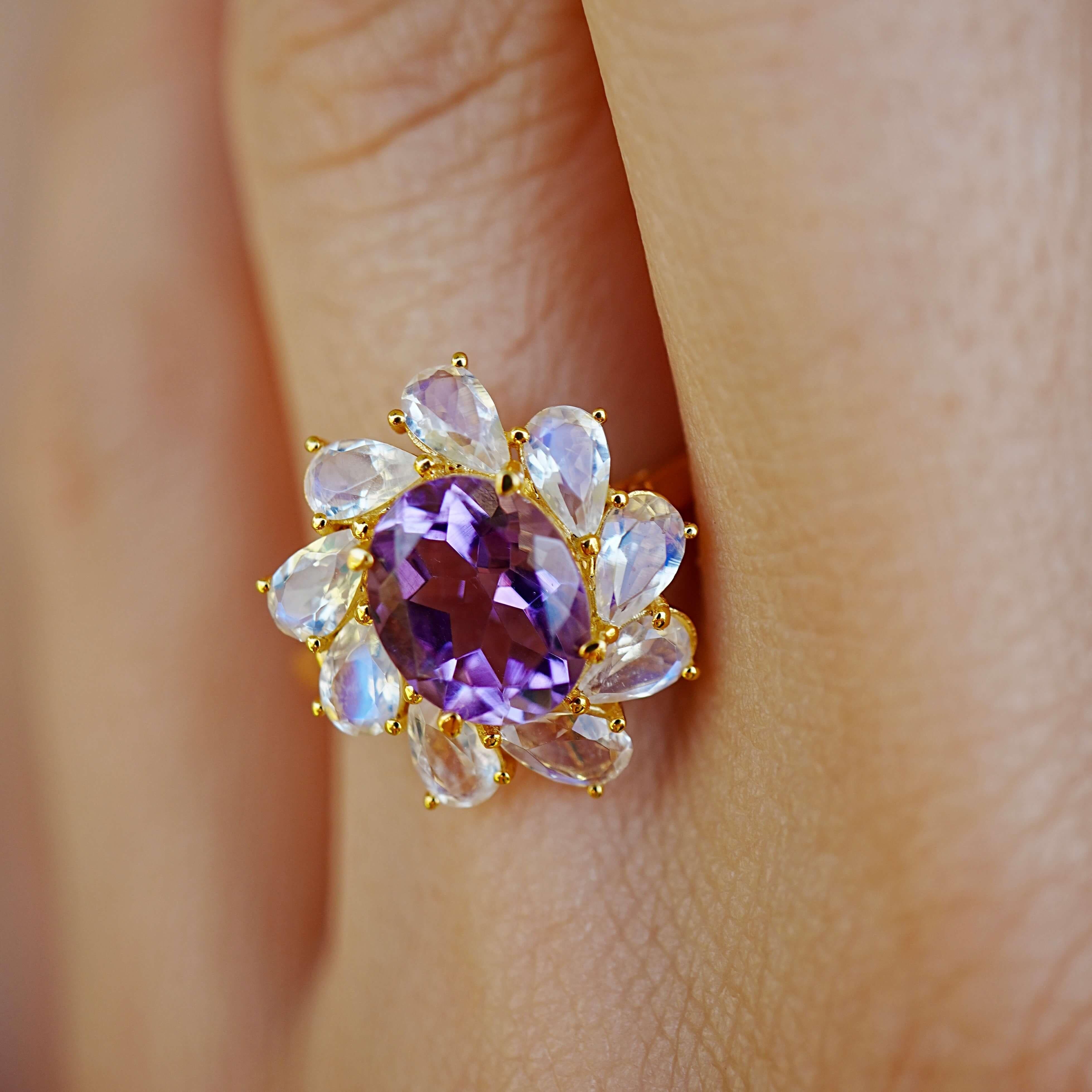 Ein Ausbruch von Violett, umrahmt von glitzerndem blauem Feuer - dieser auffällige Cocktailring zeigt einen lebhaften Amethysten in der Mitte, umringt von Regenbogen-Mondsteinen im Birnenschliff, die in sanftem, opalisierendem Licht leuchten. Das