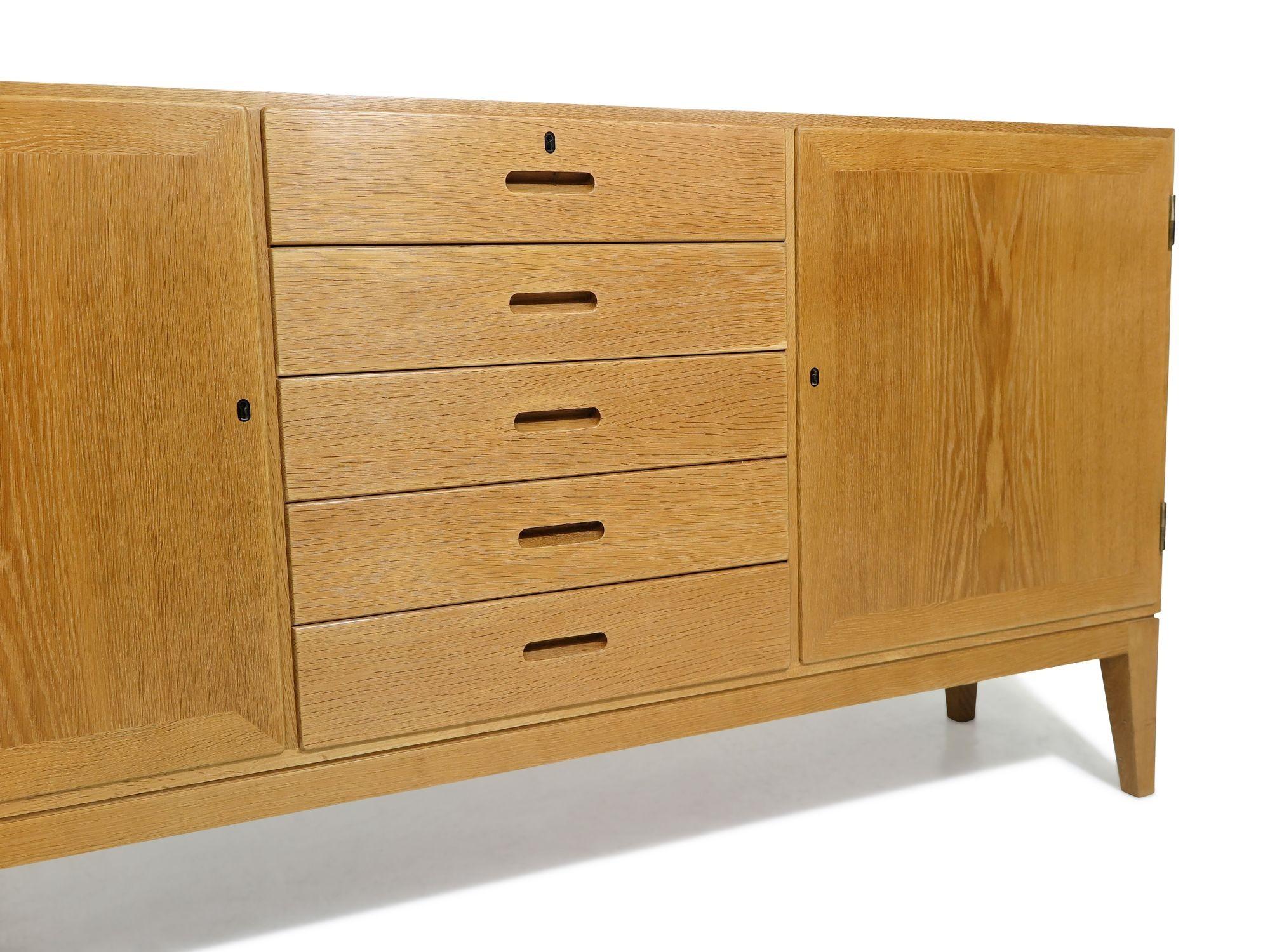 Mid-century Danish Oak credenza designed by Kai Winding in 1958, Denmark. This finely crafted cabinet showcases oak construction with mitered edges and recessed carved pulls. Features two locking doors and five drawers in the center, with an