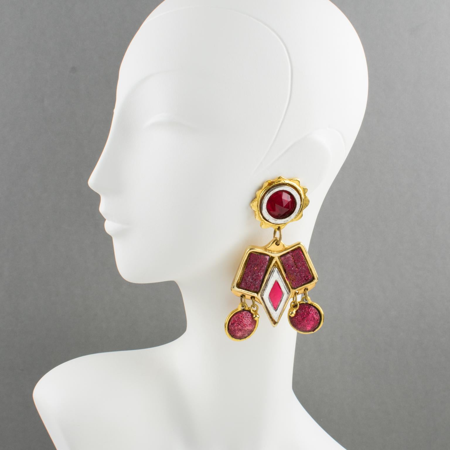 These fabulous Kalinger Paris oversized clip-on earrings feature a geometric dangling shape. The gilded-metal and silvered-metal-coated resin frame has dangling charms. All elements are ornate with ruby-red resin rhinestones and cabochons, with some