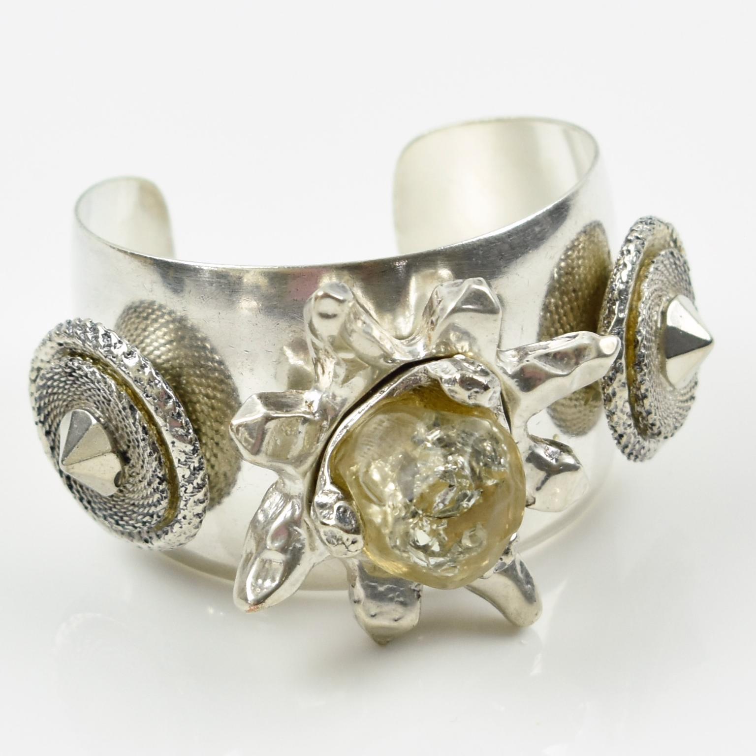 This exquisite modernist bracelet, designed by Kalinger in Paris, features a large silver-plated metal cuff bangle shape topped with three charm elements made of silver-plated metal, featuring carved wheels and a transparent resin flower. The