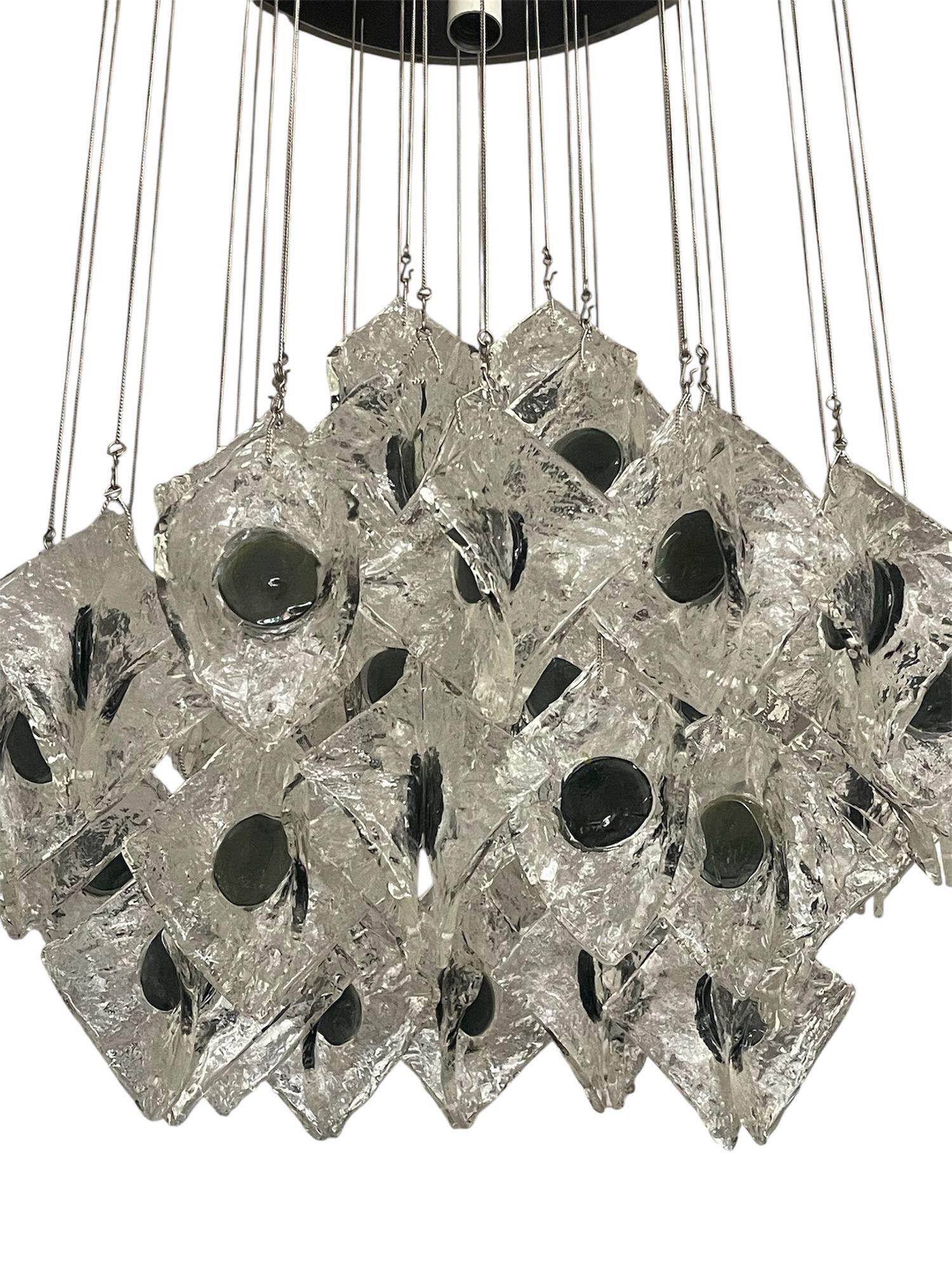 Un lustre ou un affleurant du milieu du siècle conçu par Kalmar, avec des éléments en verre fabriqués en collaboration avec Mazzega. Le luminaire se compose d'une plaque de plafond circulaire en métal noir qui suspend des pétales carrés incurvés en