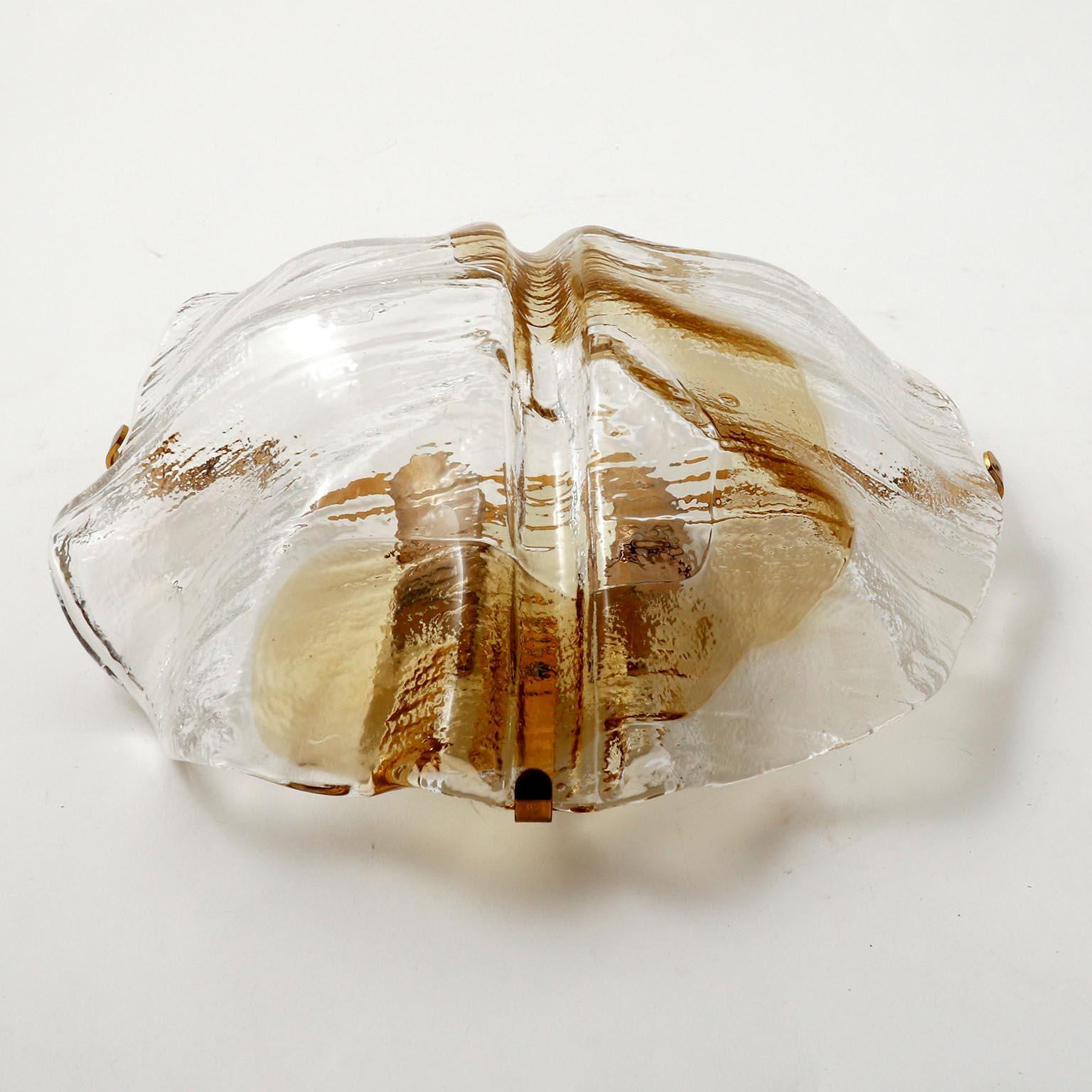 A wall light fixture by J.T. Kalmar, Austria, manufactured in midcentury, circa 1970 (late 1960s or early 1970s). 
An organic shaped clear and amber, golden or bronze toned Murano glass lamp shade is held by a brass bracket on a metal backplate. The