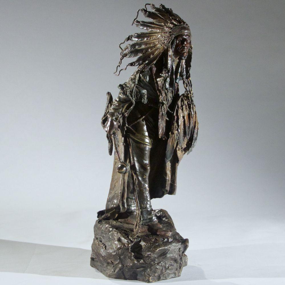 Carl Kauba (Austrian, 1865-1922), late 19th-early 20th century bronze figure of a Sioux Chief attached to bronze base, standing in war dress with a feathered shield and headdress and holding a scull cracker in his hand, stamped on the sculpture and