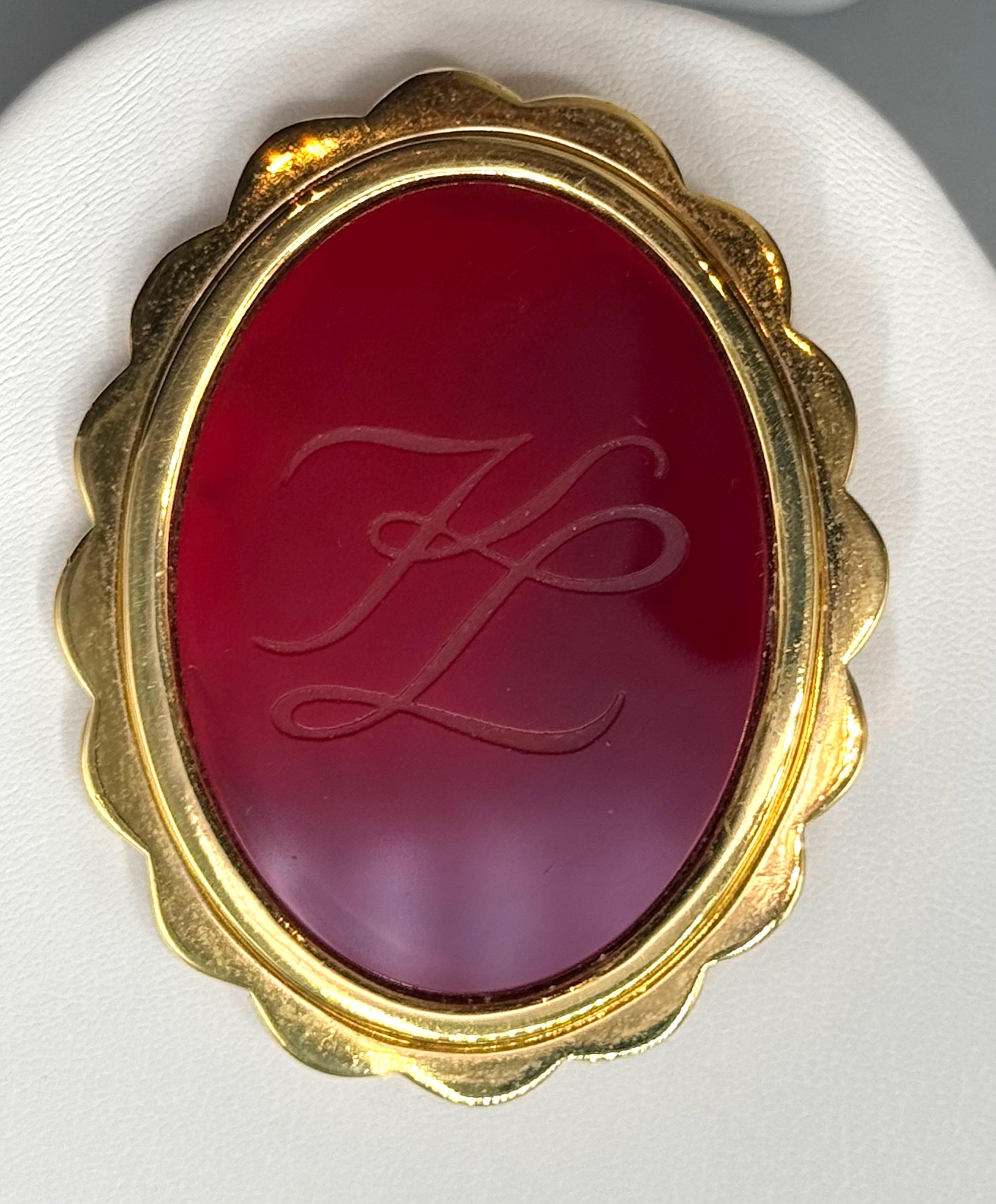 Une belle, grande et rare broche sculptée de Karl Lagerfeld datant des années 1980/90. La broche présente une grande pièce de verre ovale rouge bordeaux gravée du célèbre logo des initiales K et L entrelacées et encadrée dans une monture festonnée