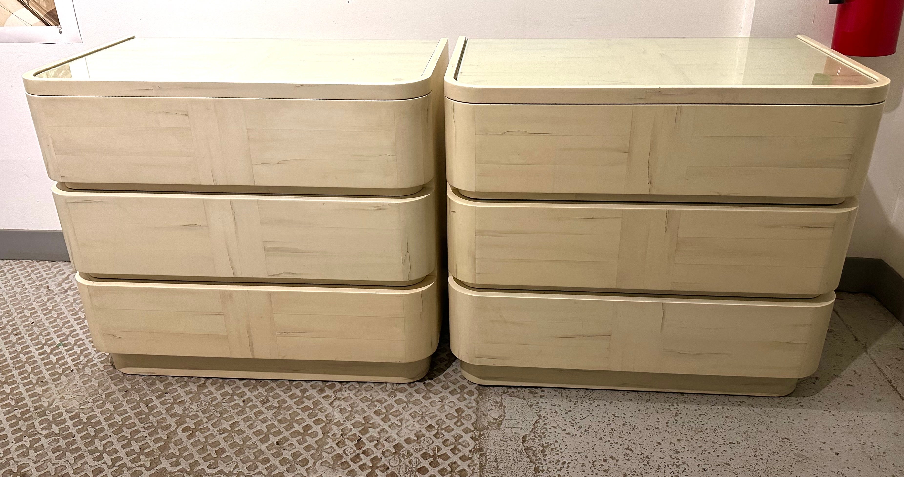 An exceptional and possibly unique pair of Karl Springer commodes with inset glass tops over three rounded drawers on an inset plinth.  What sets this set apart is the beautiful hand applied cream-lacquered finish in a trompe l'oeil antiqued
