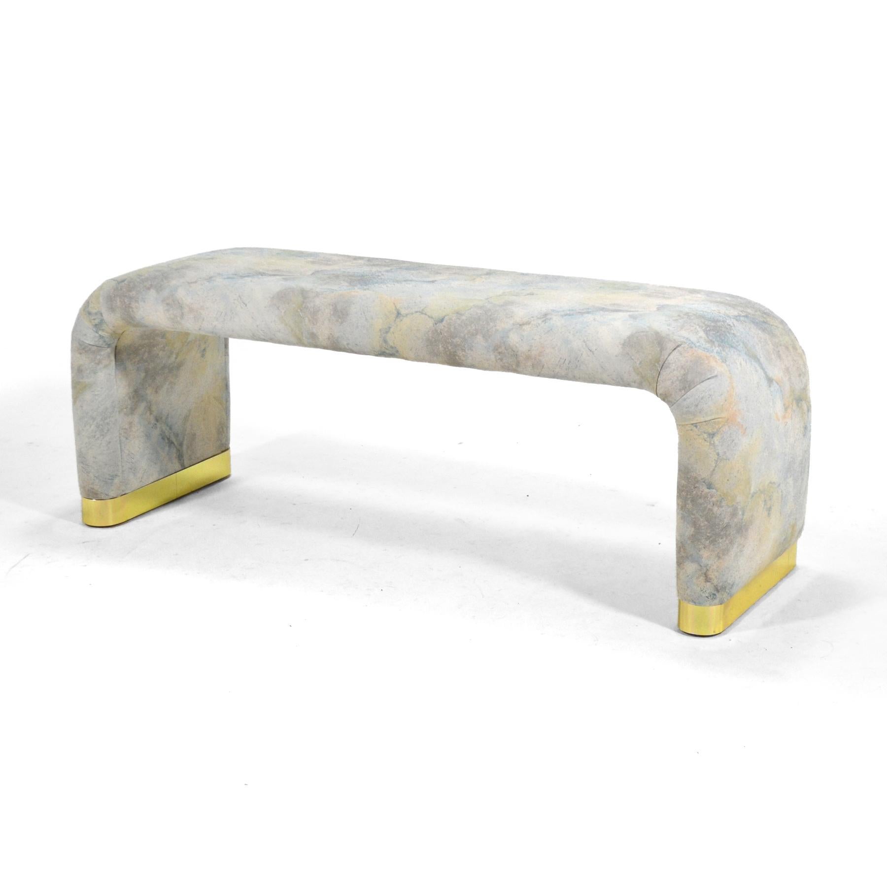 A spare, minimal design, this bench is very similar in aesthetic to the work of Karl Springer. The simple upholstered form terminates at the floor with brass feet. It currently retains its original 1980s upholstery, but will look spectacular once it